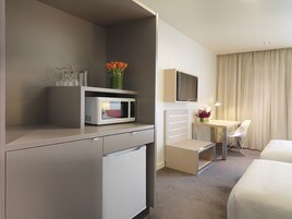 Quarto de hotel com cozinha compacta, cama, escrivaninha com cadeira, televisão e janela com cortinas.