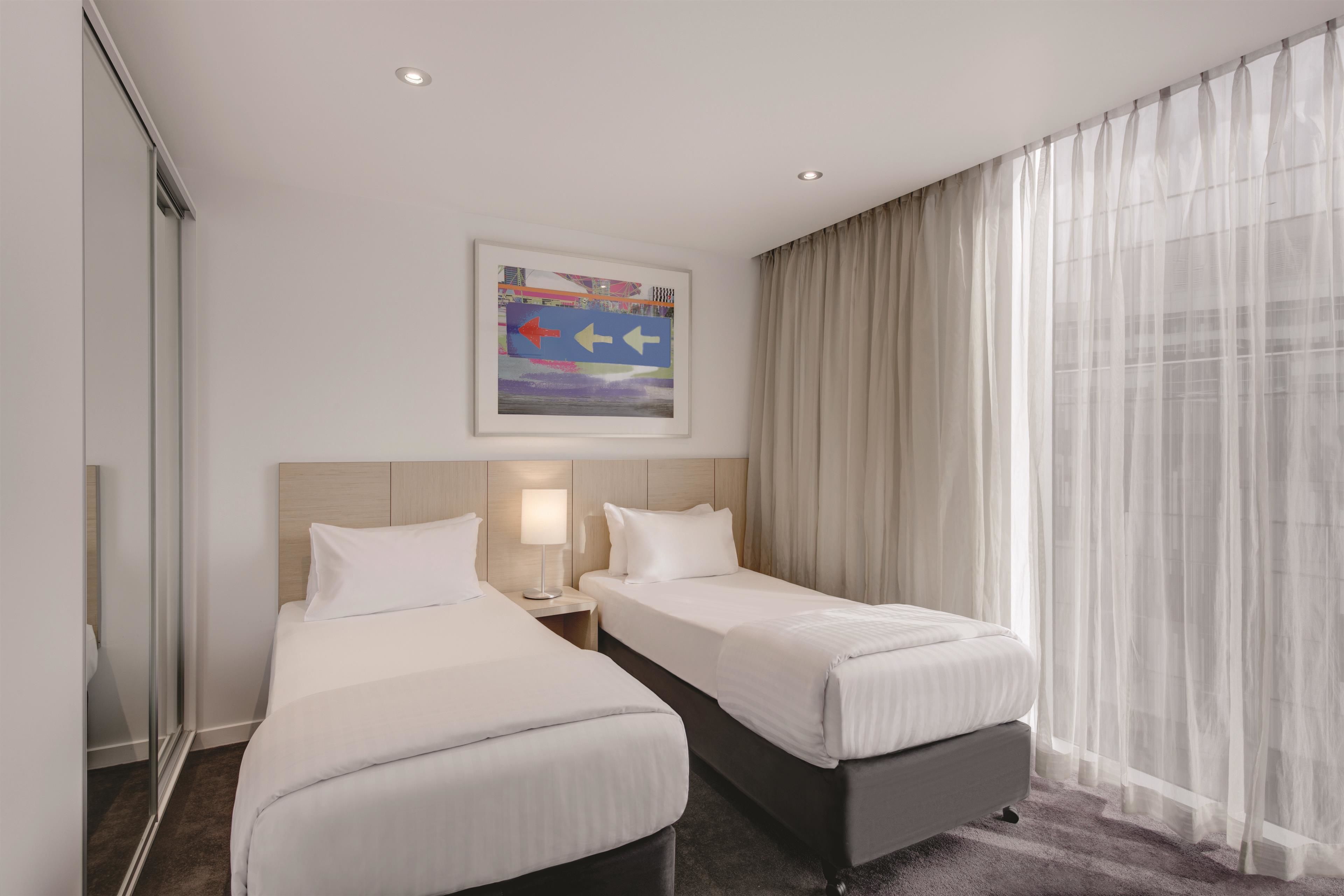 Habitación de hotel con dos camas, una mesita de noche, una lámpara, un cuadro en la pared y cortinas translúcidas.