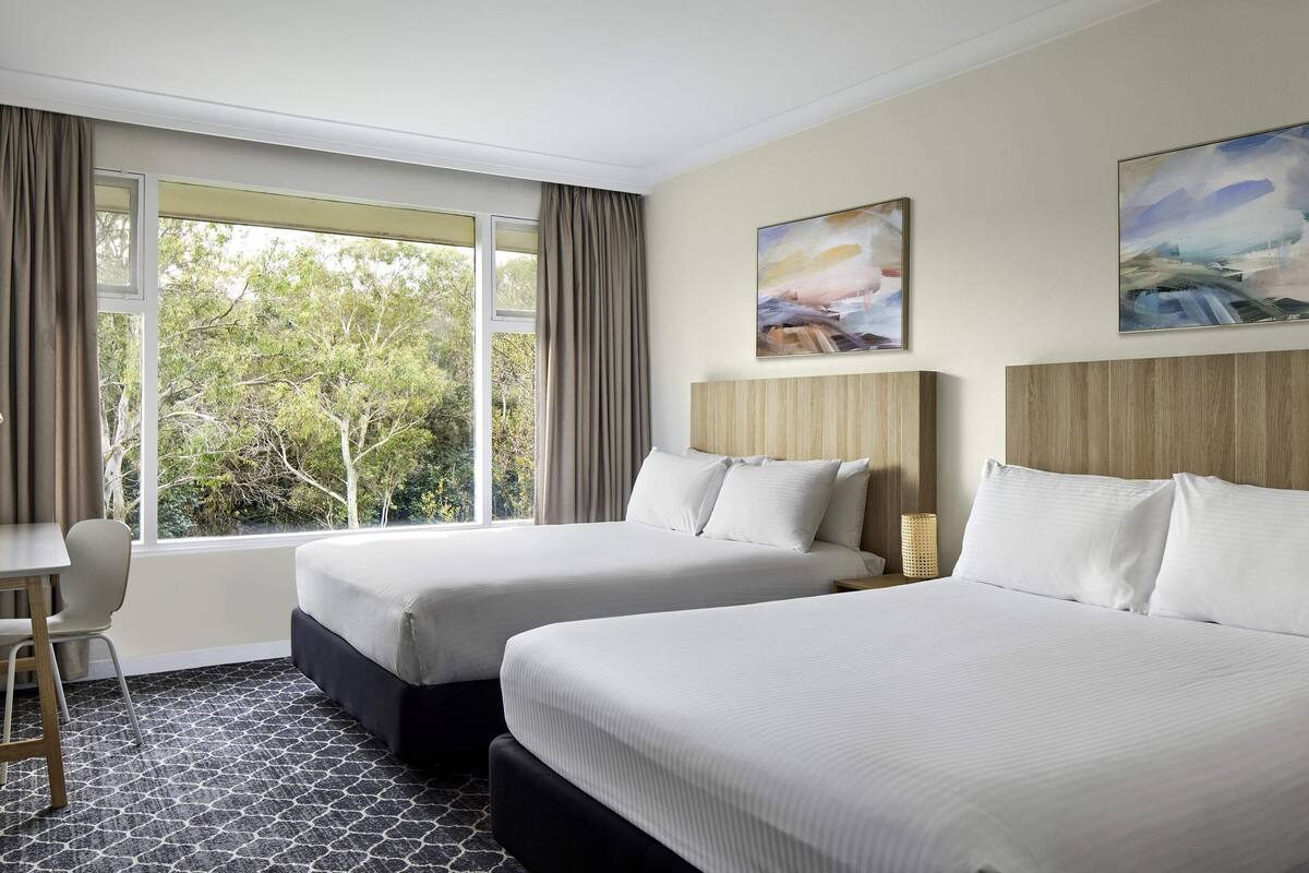A hotel room with two beds, a desk, and a view of trees through large windows.