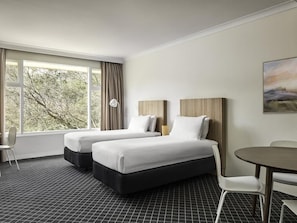 A hotel room with two beds, a dining table, a chair, and a large window with curtains.