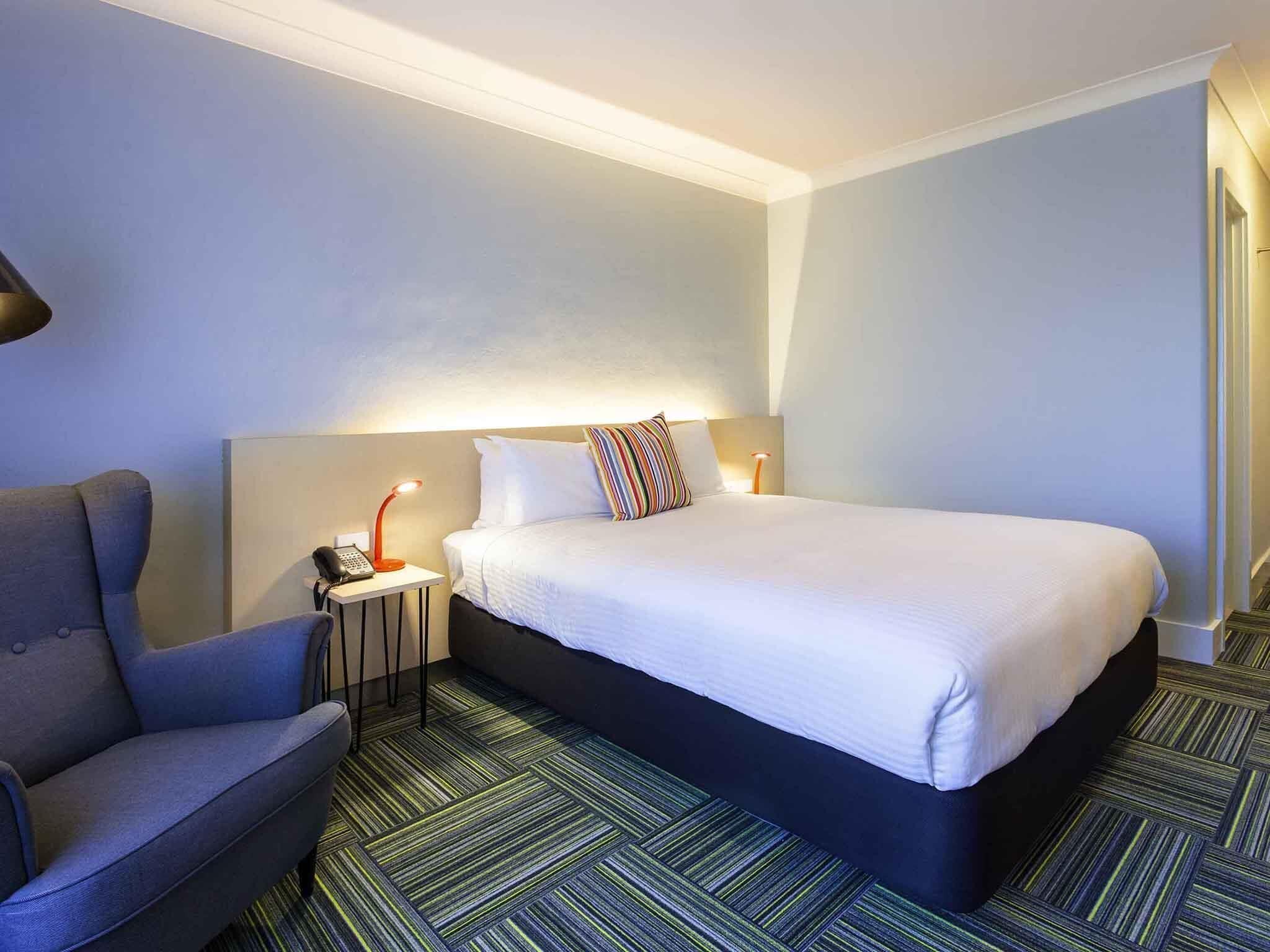 Een hotelkamer met een bed, een stoel, een nachtkastje met een lamp en een kamerbreed tapijt met dessin.
