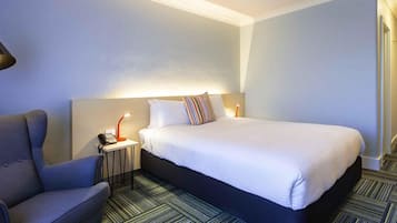 A hotel room with a bed, a chair, a nightstand with a lamp, and a patterned carpet.