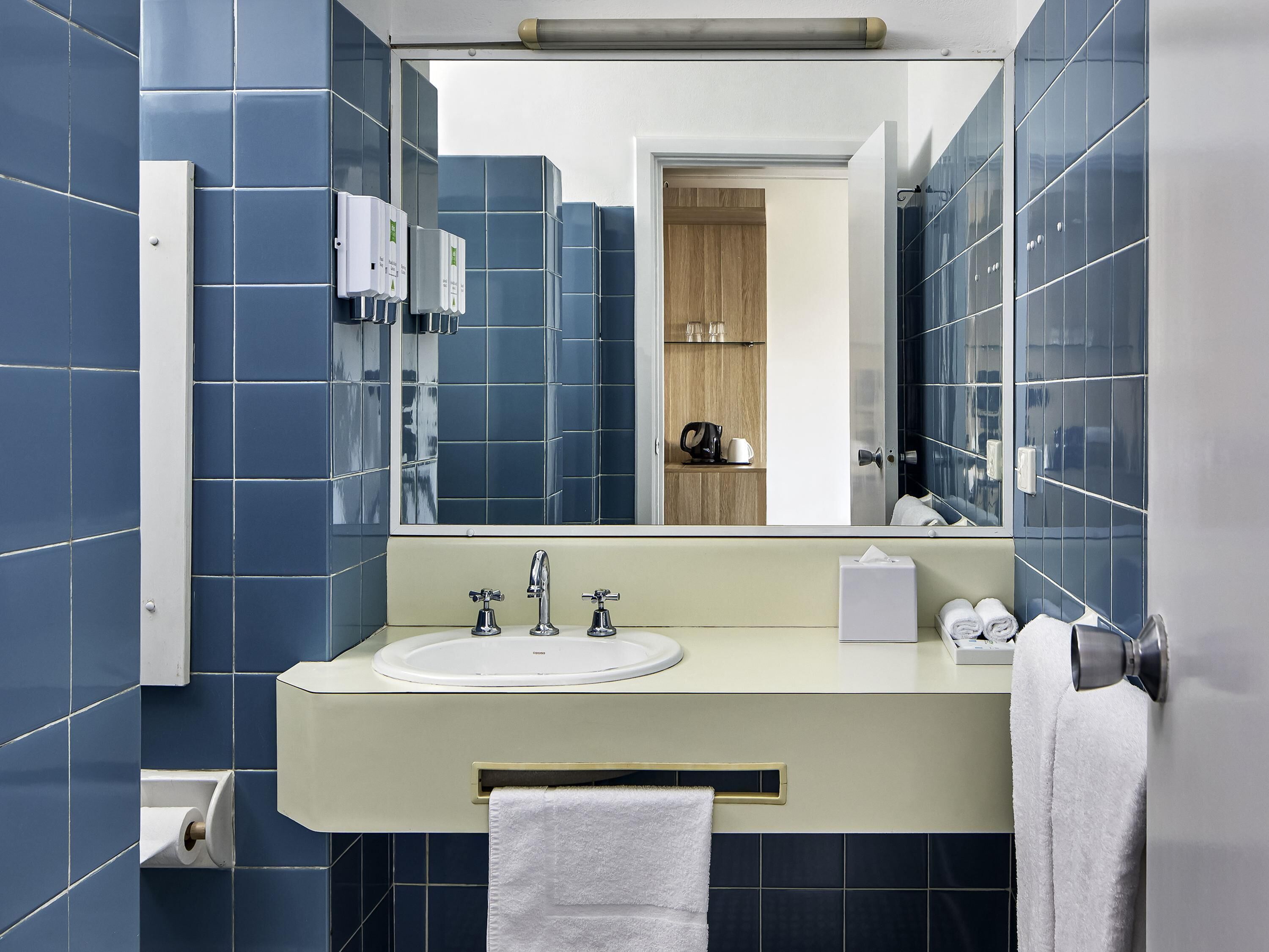 A modern bathroom with blue tiles, a beige sink, and a mirror reflecting a wooden door and shelves.