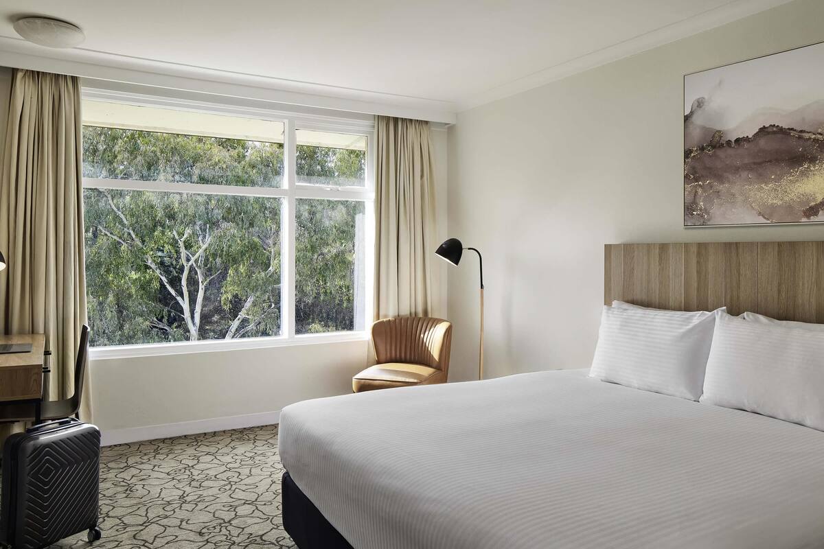A hotel room with a large bed, a desk, a chair, a suitcase, and a window with curtains.