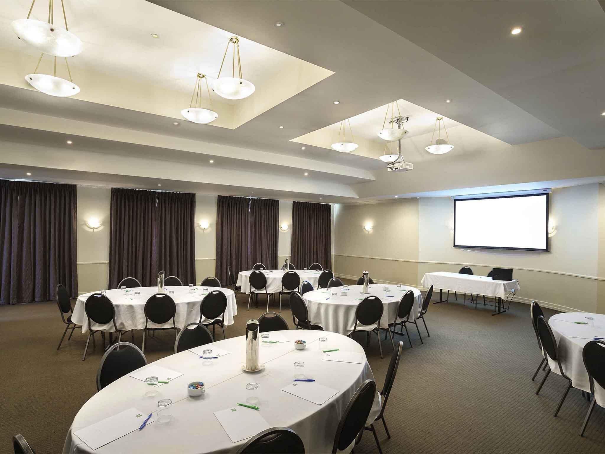 Una sala conferenze con tavoli rotondi, sedie, uno schermo per proiettore e luci a soffitto.