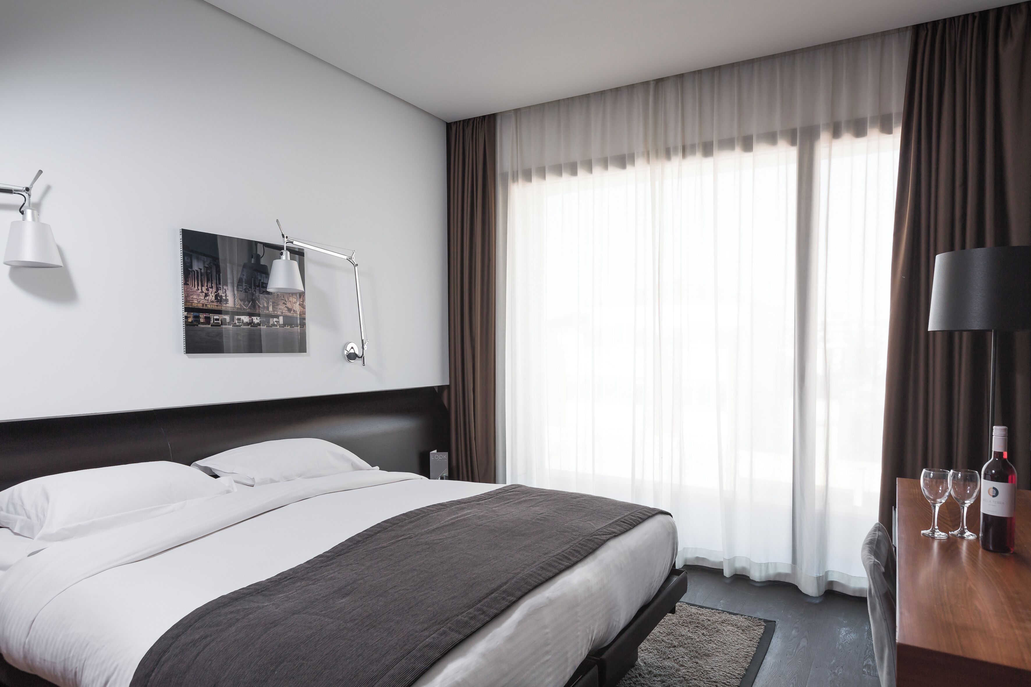 A modern hotel room with a large bed, a desk with a lamp, a wine bottle, and glasses.