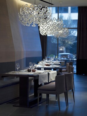 A modern dining area with a unique chandelier, tables set for dining, and a view of the outdoors through large windows. - The Met Hotel, a member of Design Hotels (Thessaloniki)