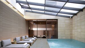 Un espace piscine intérieur moderne, doté de chaises longues en bois, d’une salle de spa fermée par des baies vitrées et offrant une vue dégagée sur le ciel à travers le toit.