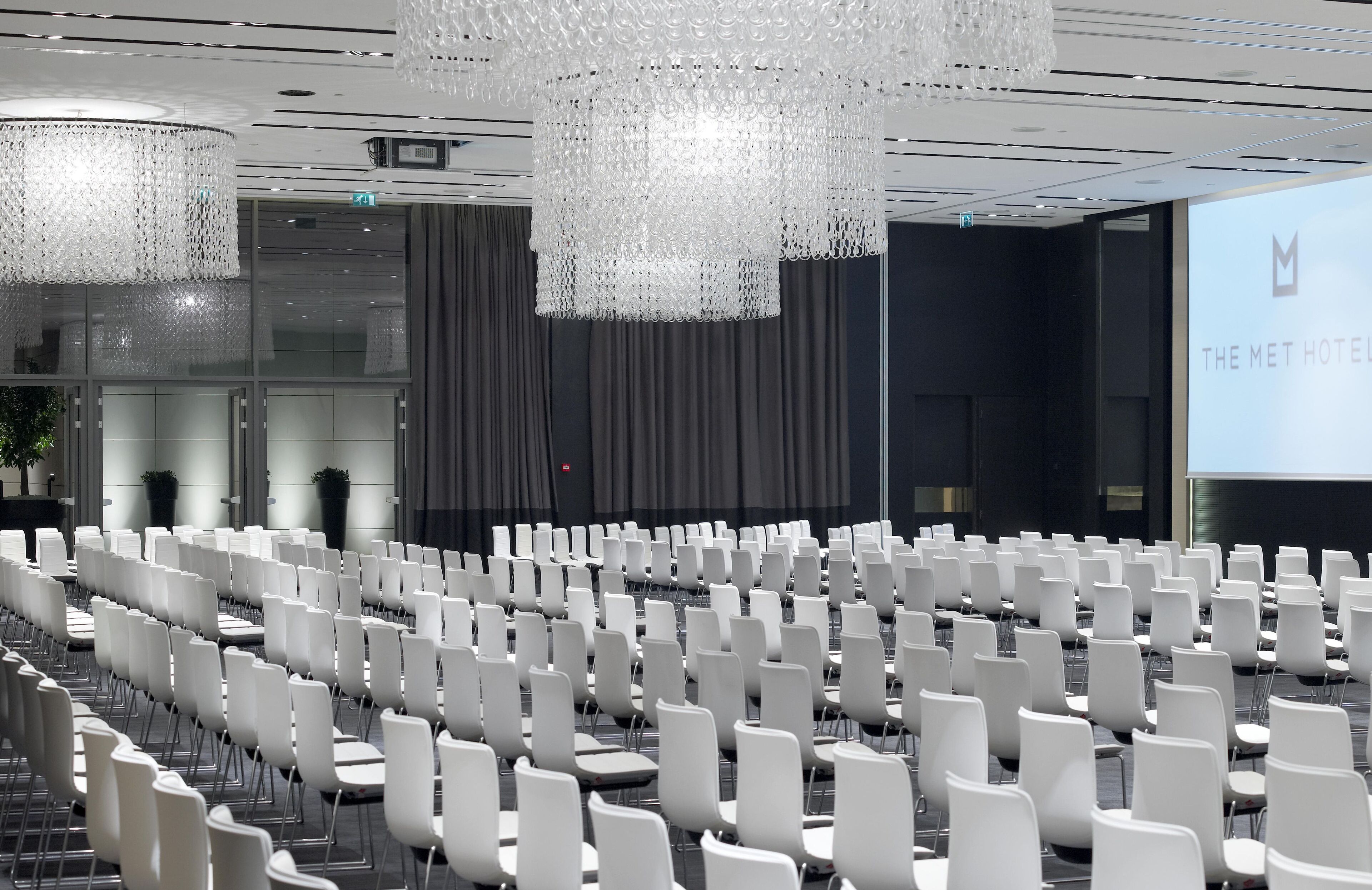 Una sala conferenze con file di sedie bianche, un grande schermo che mostra 'THE MET HOTEL' e un lampadario moderno.