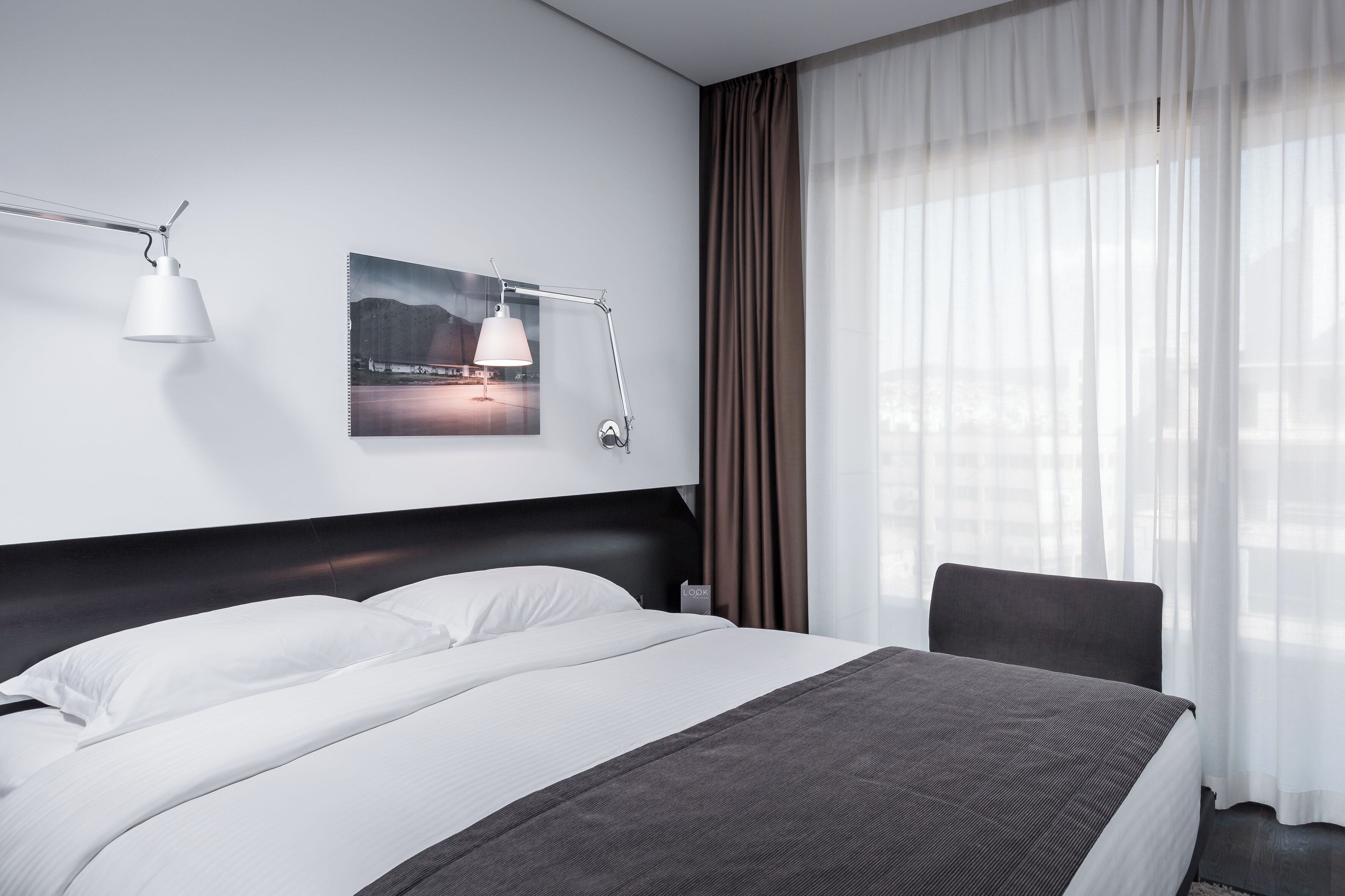 A modern hotel room with a large bed, a desk lamp, a picture on the wall, and a view of the outdoors through sheer curtains.
