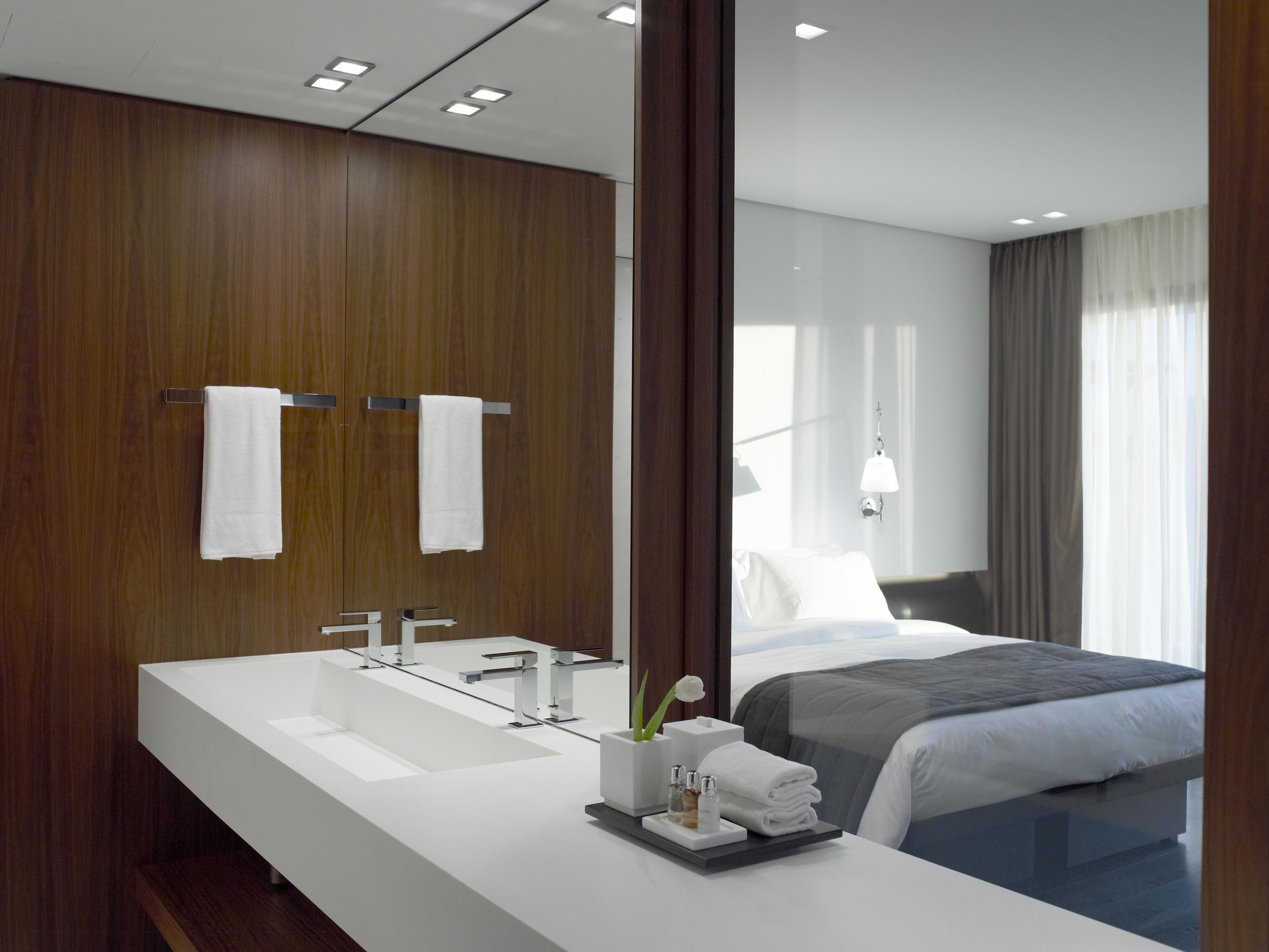 A modern hotel bathroom with a large white sink, two towel racks, and a mirror reflecting a bedroom with a bed and a window.