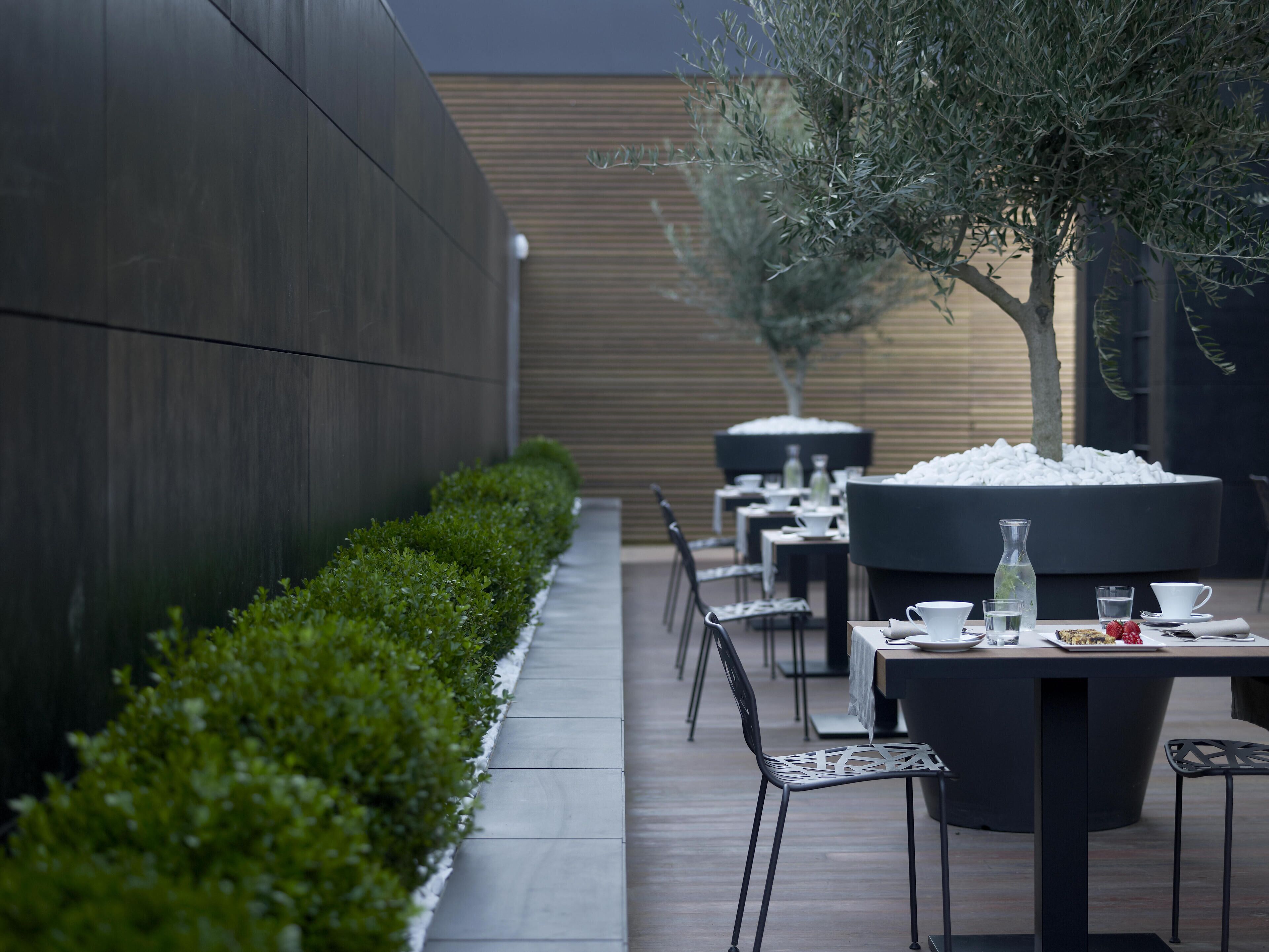 An outdoor dining area with tables and chairs, a pathway, and a row of bushes along the wall.
