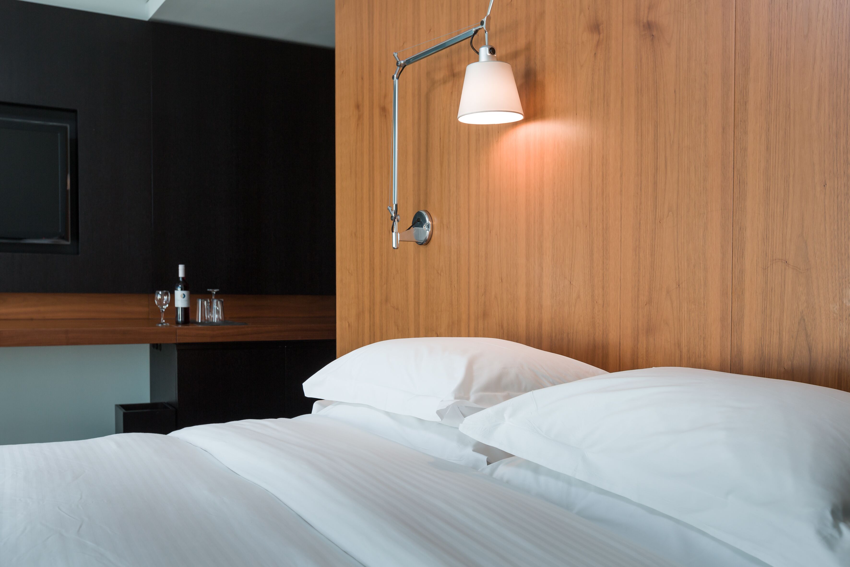 A modern hotel room with a wooden headboard, a bedside table with a lamp, and a neatly made bed.