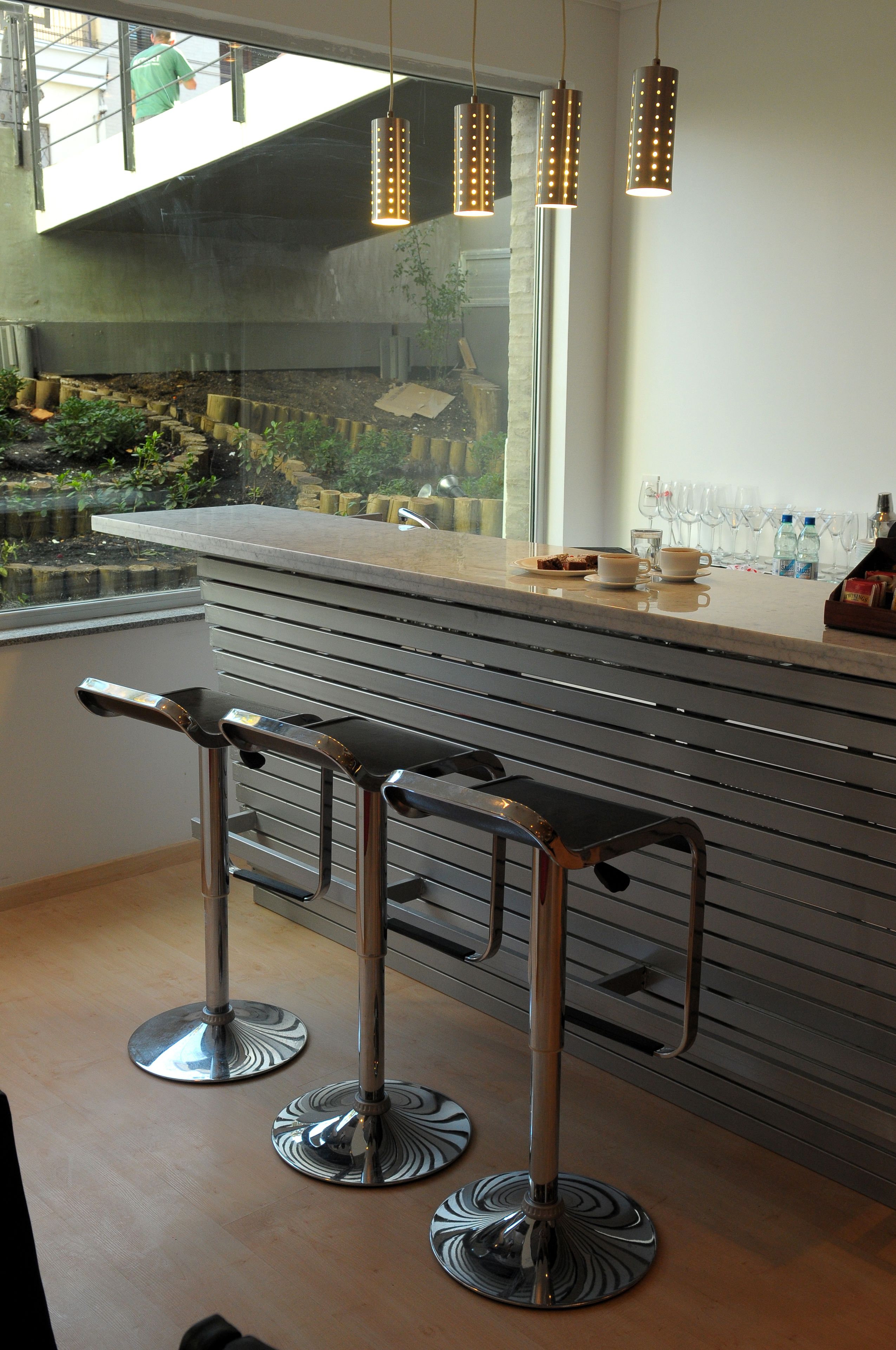Zona de bar moderna con dos taburetes, una barra con vitrina de vidrio y vistas a un jardín a través de un ventanal.