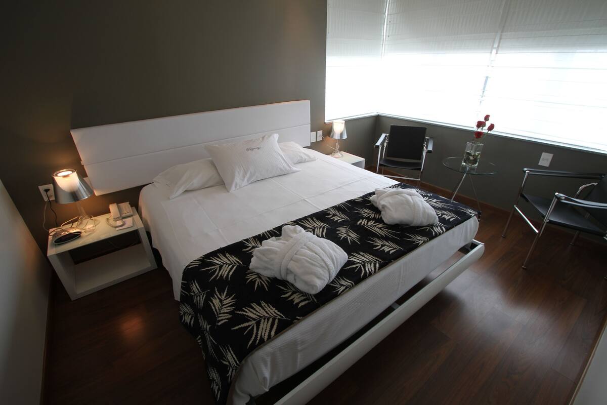 Moderni hotellihuone, jossa on suuri sänky, yöpöytä lampun kanssa, tuoli ja pieni pöytä, jossa on maljakko.