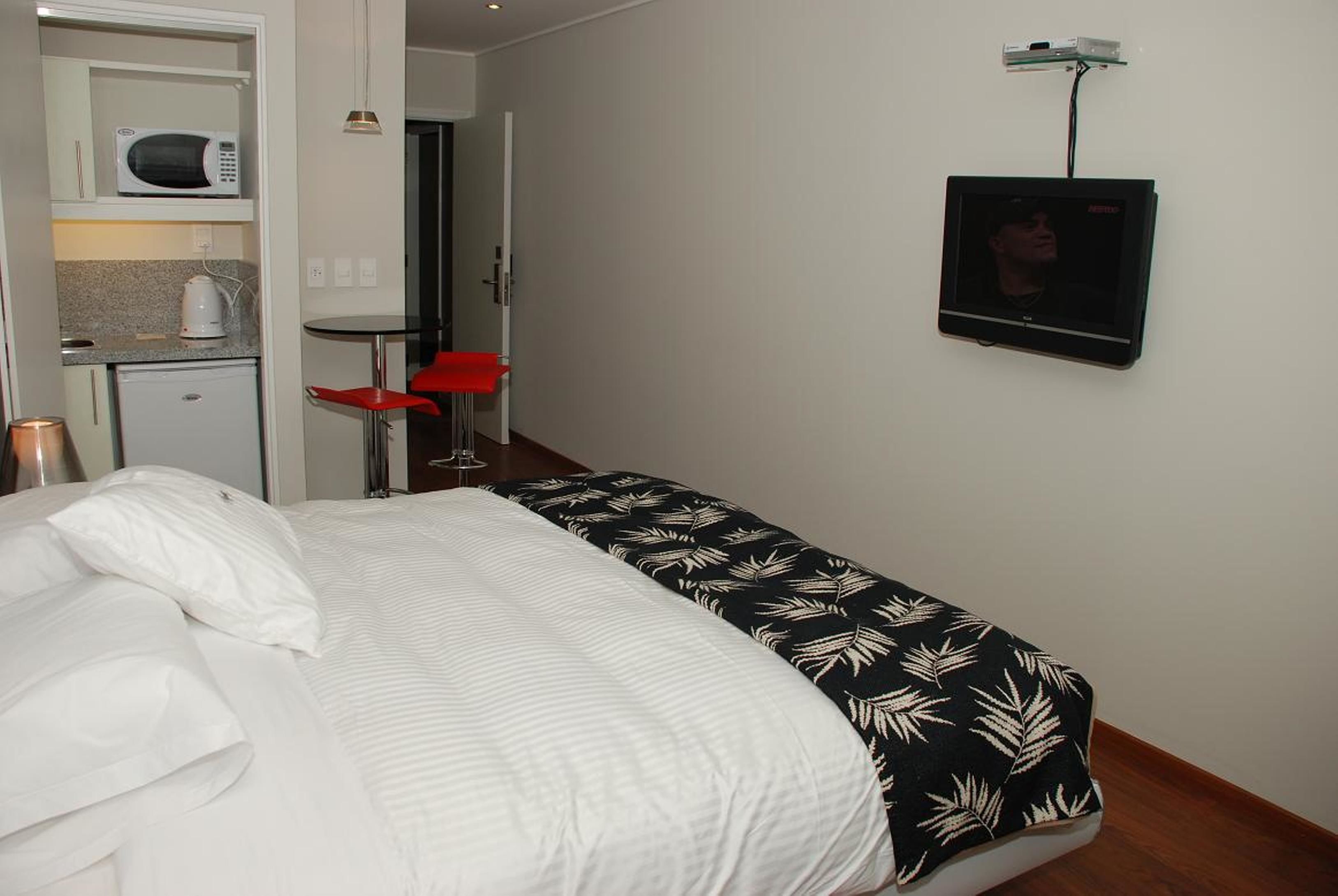 Habitación de hotel con cama, una kitchenette pequeña, televisor montado en la pared y una silla roja.