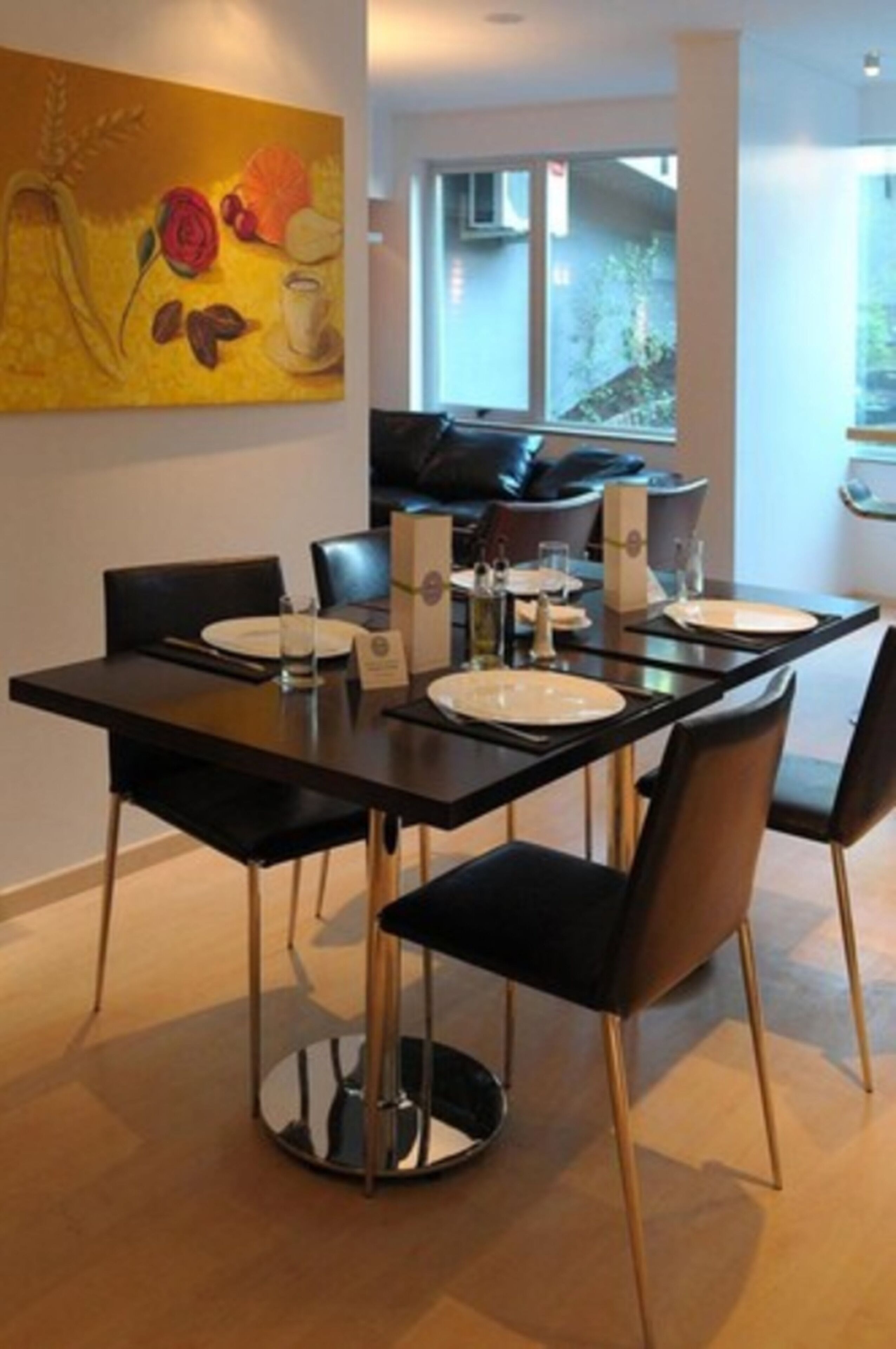 Un espace repas moderne avec une table rectangulaire dressée pour quatre personnes, des chaises noires aux pieds dorés et un tableau jaune accroché au mur.