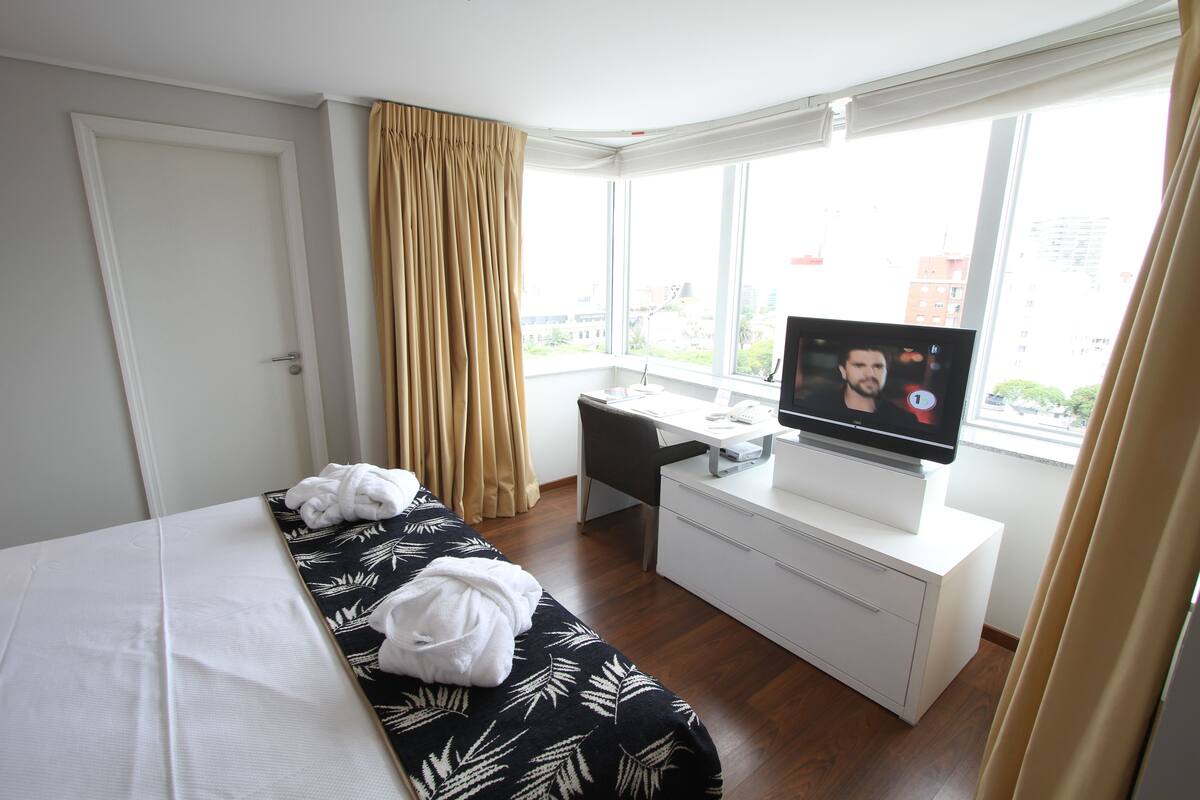 Hotellihuone, jossa on sänky, työpöytä, televisio ja ikkuna, jossa on verhot.