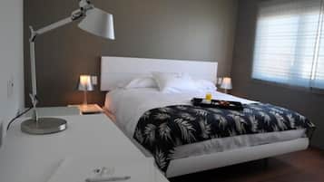 A modern bedroom with a white bed, a desk with a lamp, and a window with blinds.
