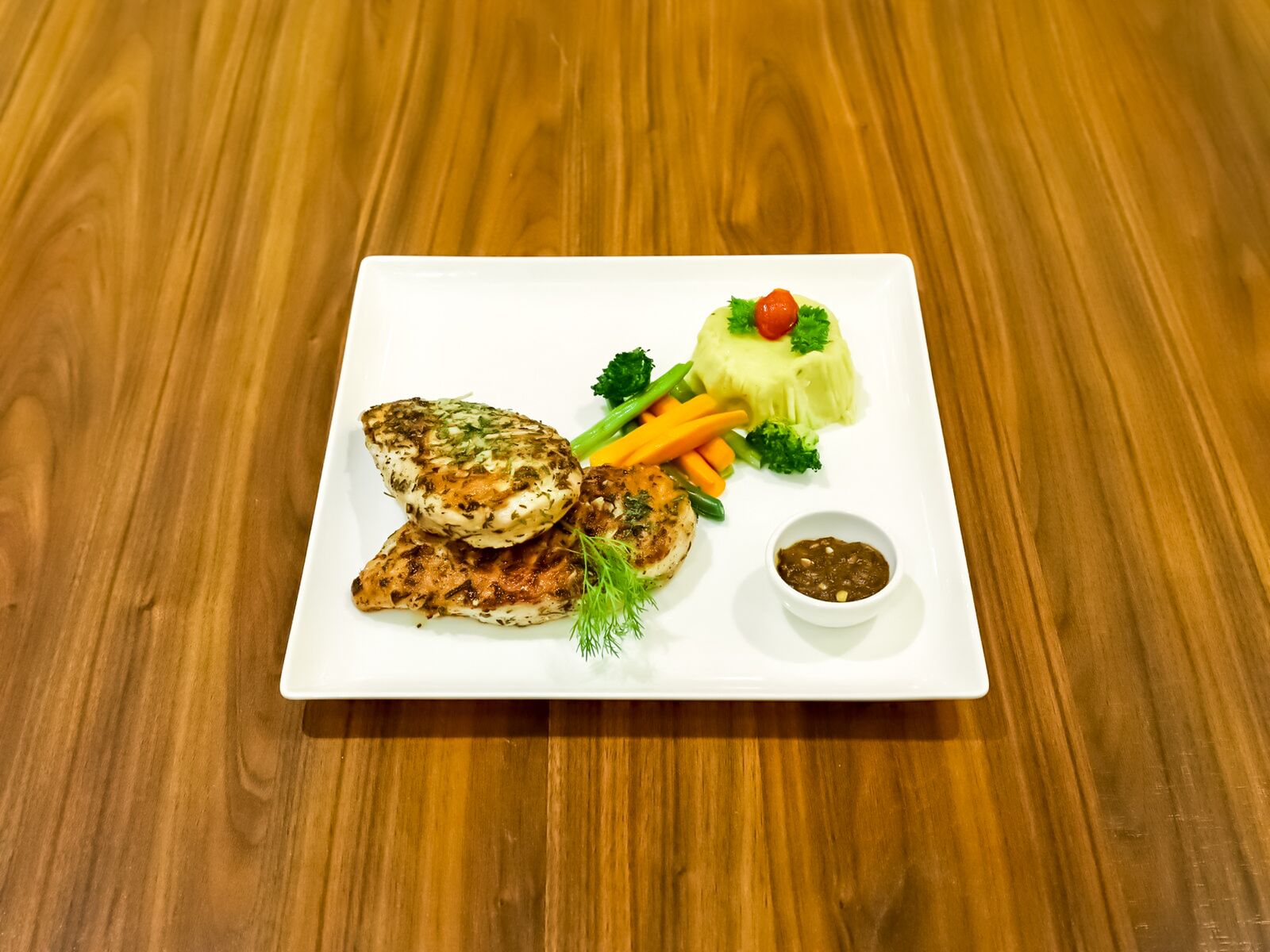 A white rectangular plate with grilled chicken, carrots, broccoli, and a small bowl of sauce on a wooden table.