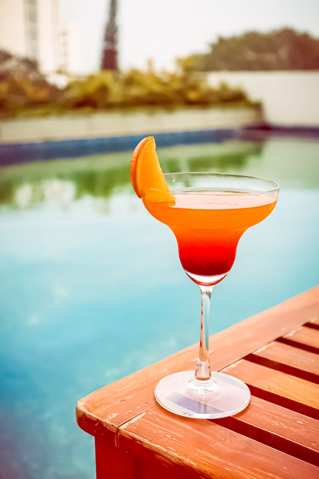 Un cocktail in un bicchiere da martini con una guarnizione d'arancia su un tavolo di legno vicino a una piscina.