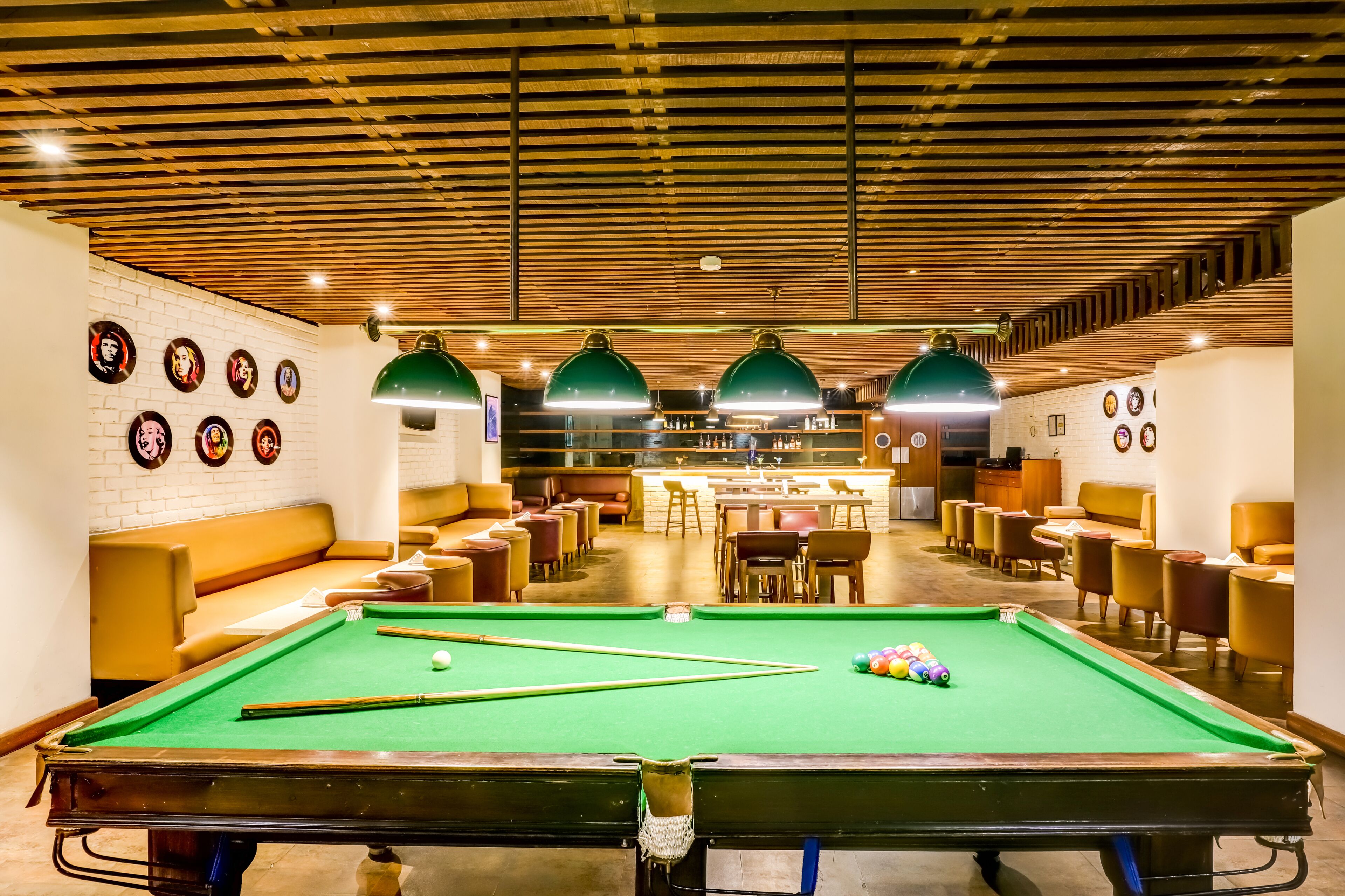 A pool table with green felt, surrounded by chairs and tables, in a room with wooden ceiling beams and wall-mounted clocks.