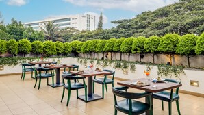 An outdoor dining area with tables and chairs, surrounded by lush greenery and a view of a modern building. - Lemon Tree Hotel, Electronics City - Bengaluru (Bengaluru)