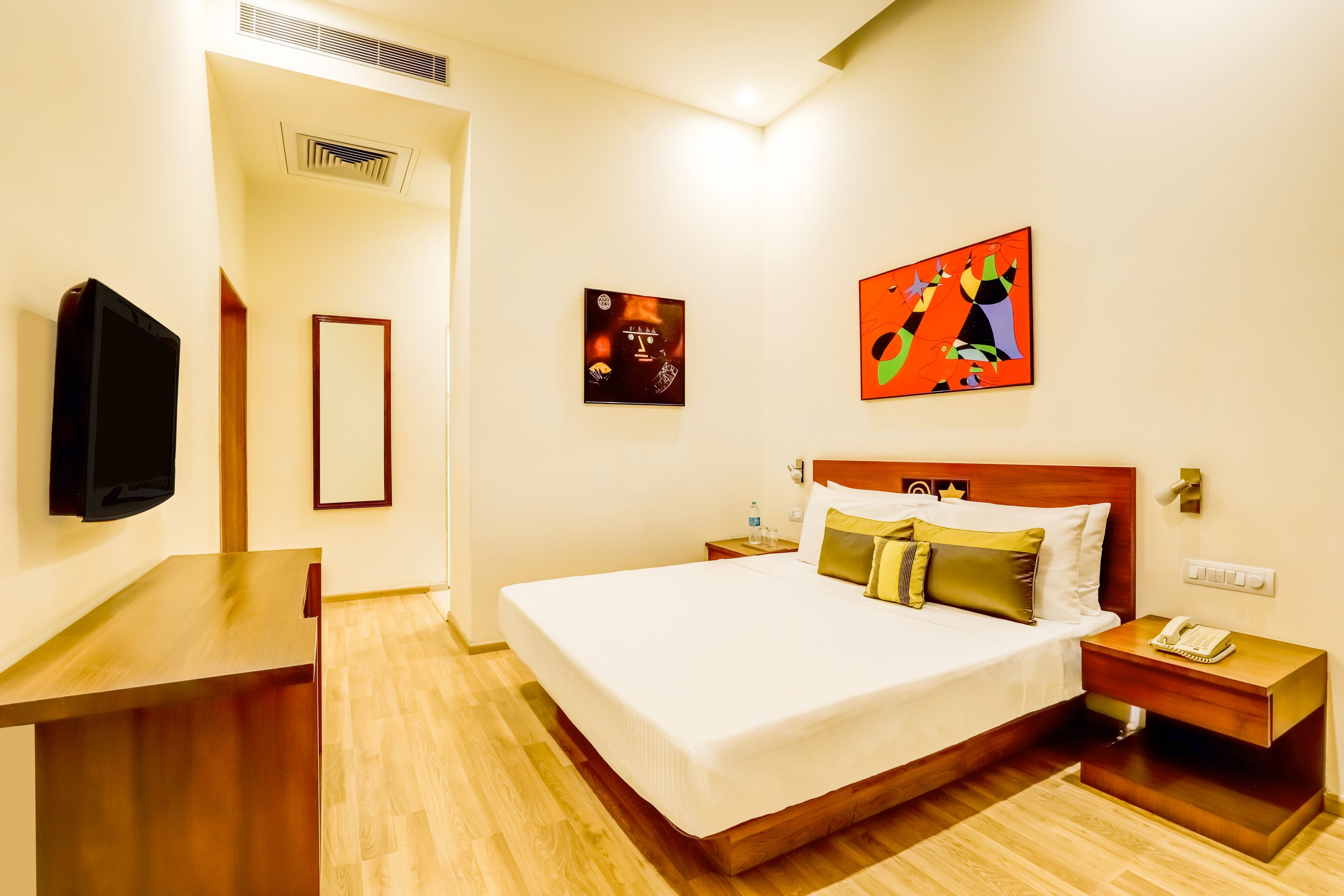 Une chambre d’hôtel avec un grand lit, une table de chevet, une commode, un téléviseur fixé au mur et deux tableaux.