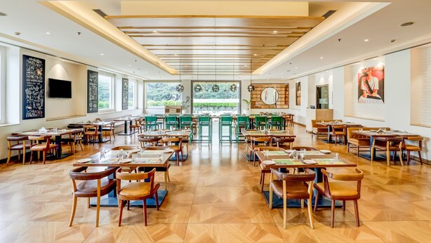 A spacious restaurant with wooden flooring, green upholstered chairs, and a central seating area. - Lemon Tree Hotel, Electronics City - Bengaluru (Bengaluru)