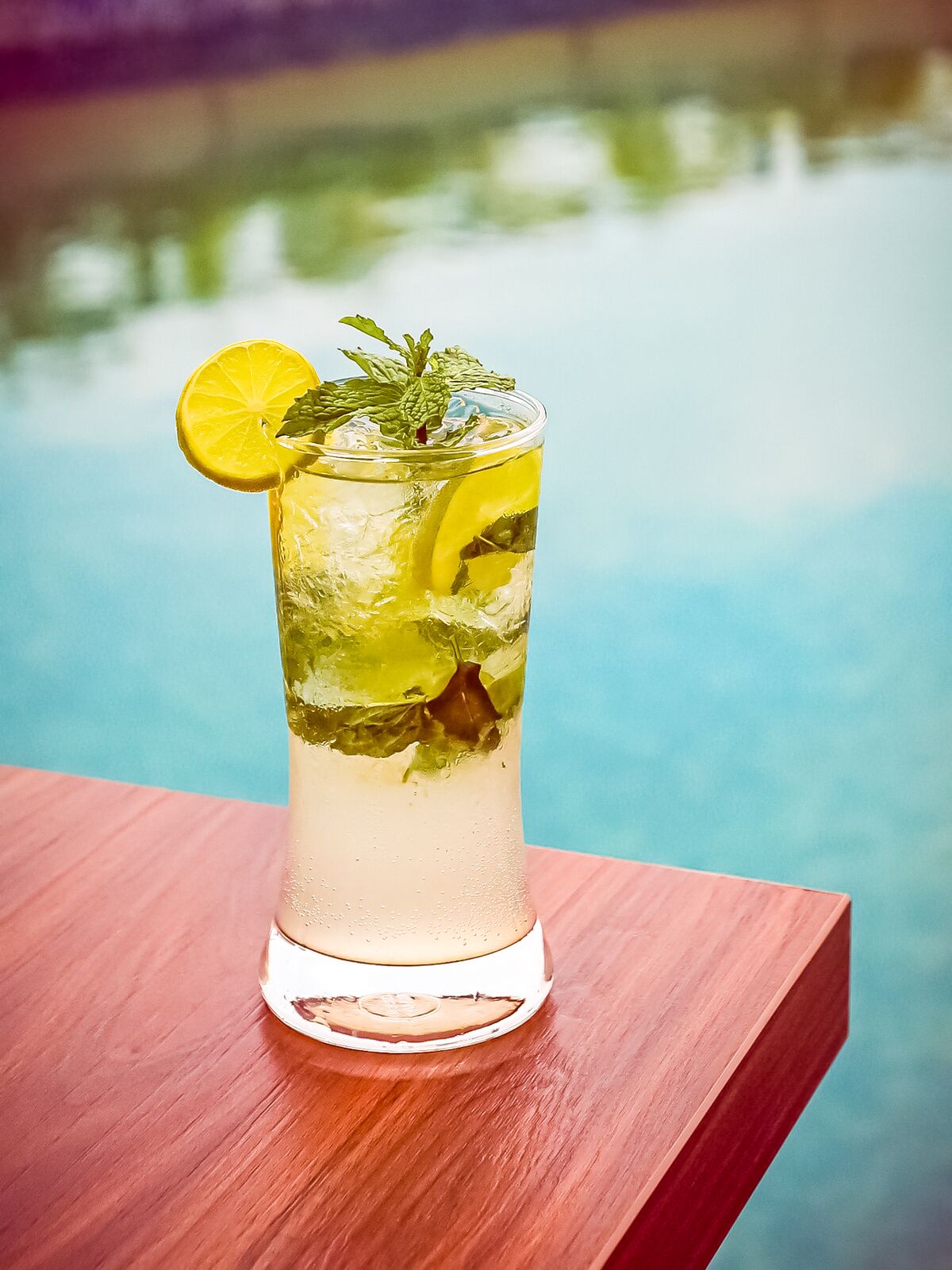 A tall glass with a clear liquid, ice, lemon slices, and mint leaves, placed on a wooden surface with a pool in the background.
