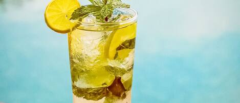 A tall glass with a clear liquid, ice, lemon slices, and mint leaves, placed on a wooden surface with a pool in the background.