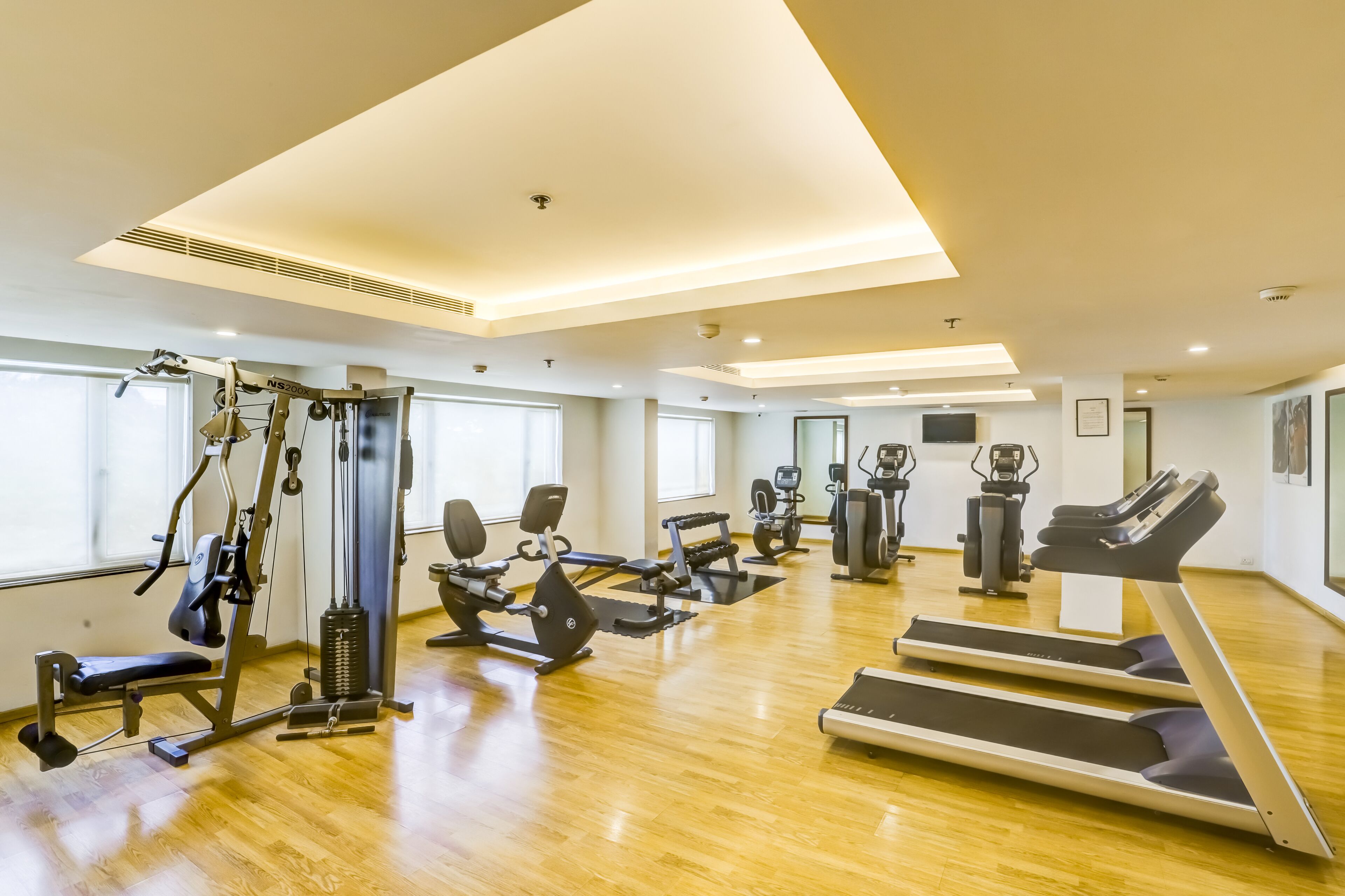 Un gimnasio amplio con diversos equipos de ejercicio, ventanas grandes y suelo de madera.