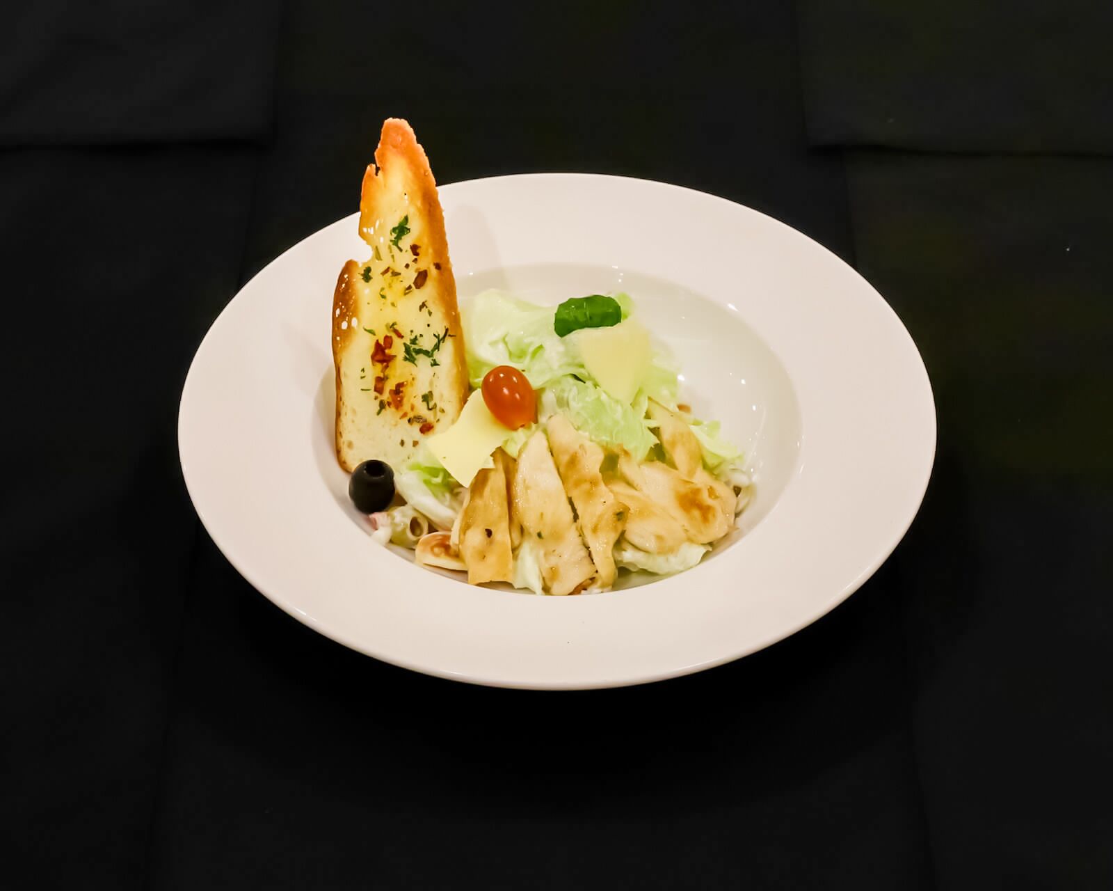 A white plate with a salad, including lettuce, cherry tomatoes, and a piece of grilled bread, on a black tablecloth.