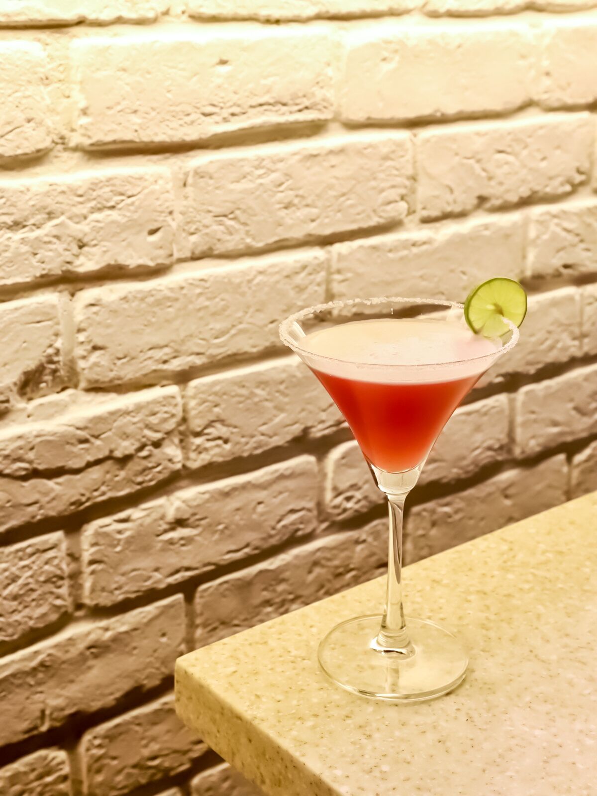 Un verre à martini, garni d’une tranche de citron vert, posé sur un comptoir à motifs, avec en arrière-plan un mur de briques blanches à la texture marquée.