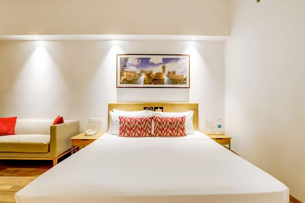 Une chambre d’hôtel avec un grand lit, une table de chevet avec un téléphone, une autre table de chevet avec une bouteille d’eau, un oreiller rouge et blanc à motifs, et un tableau encadré au mur.