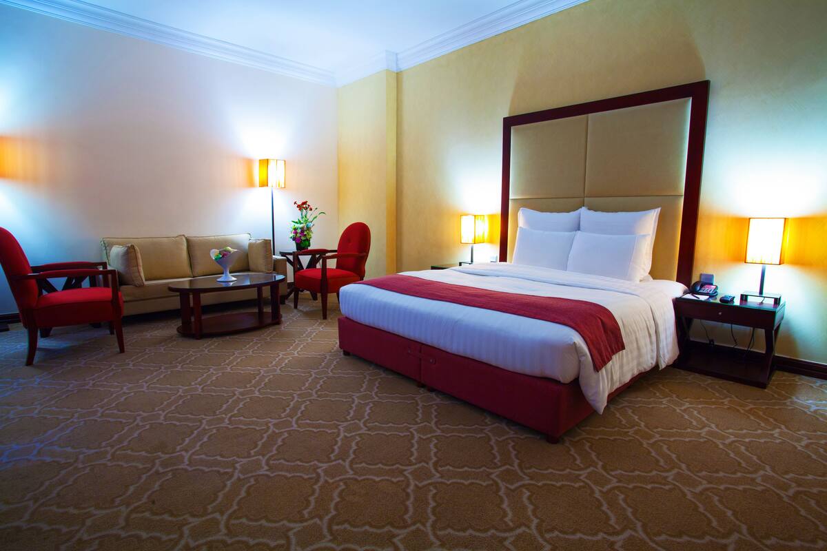 A hotel room with a large bed, a sofa, a red armchair, a small table, and two lamps.