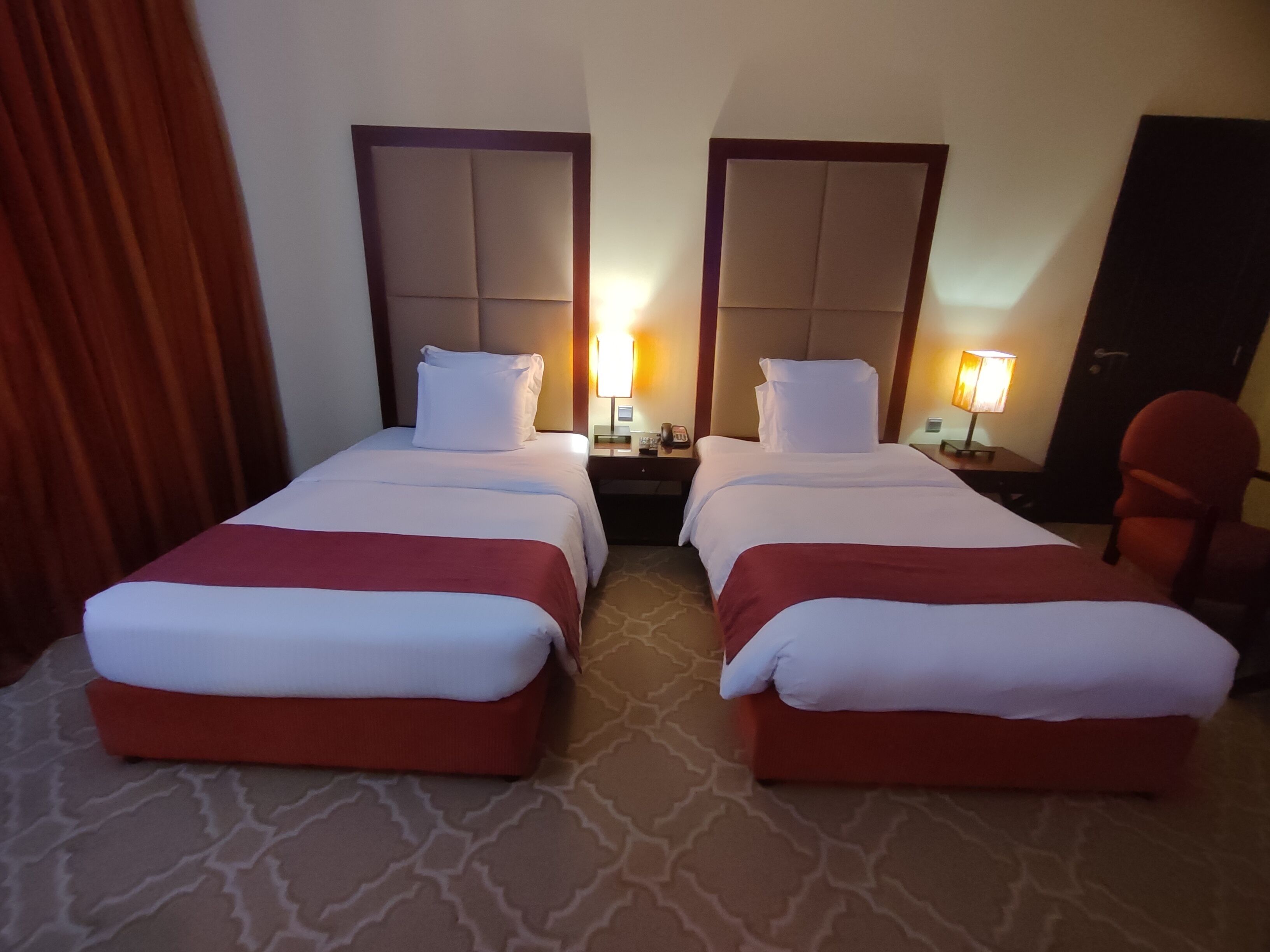 A hotel room with two beds, a red carpet, a wooden headboard, a nightstand with a lamp, and a red chair.