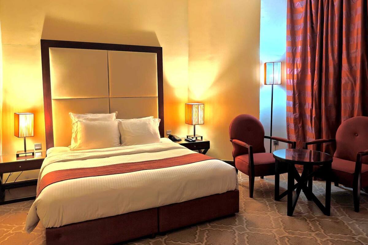 A hotel room with a large bed, two bedside lamps, a small table with chairs, and a view of the outside through a window with curtains.