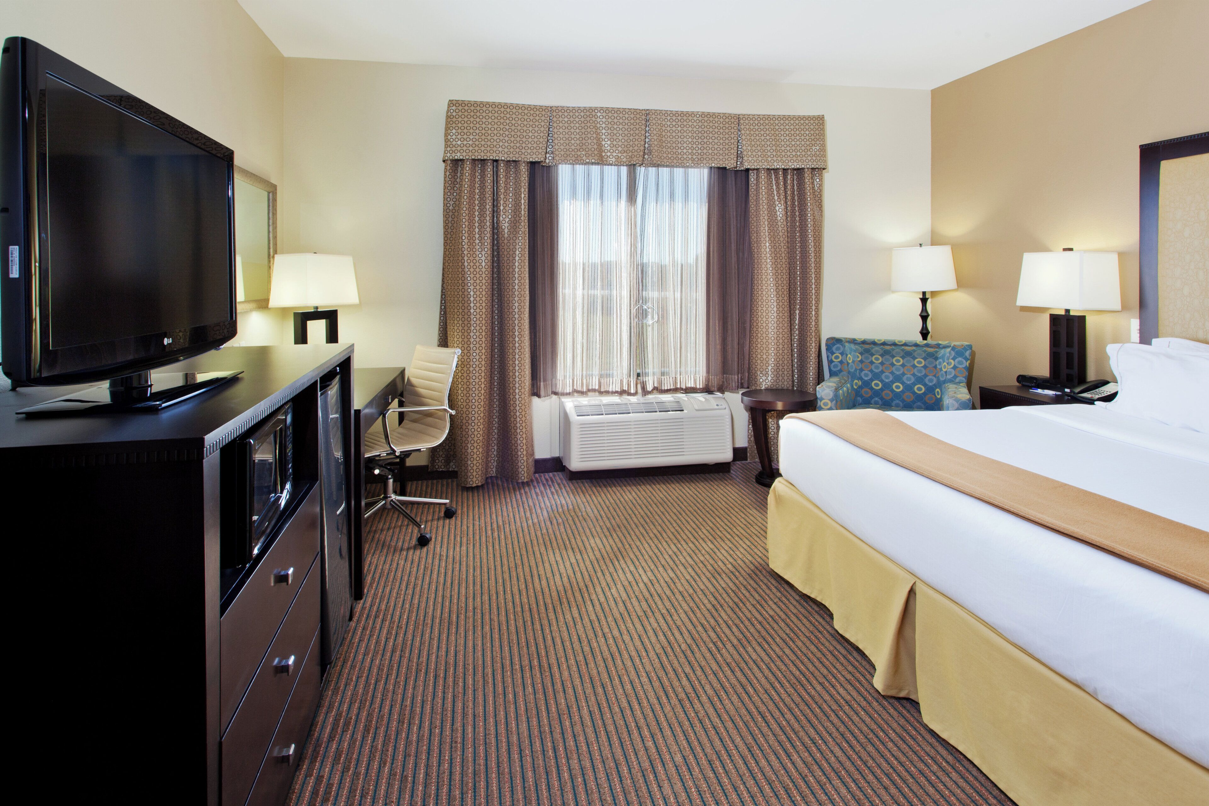 A hotel room with a bed, a TV, a desk, a chair, a lamp, and a window with curtains.