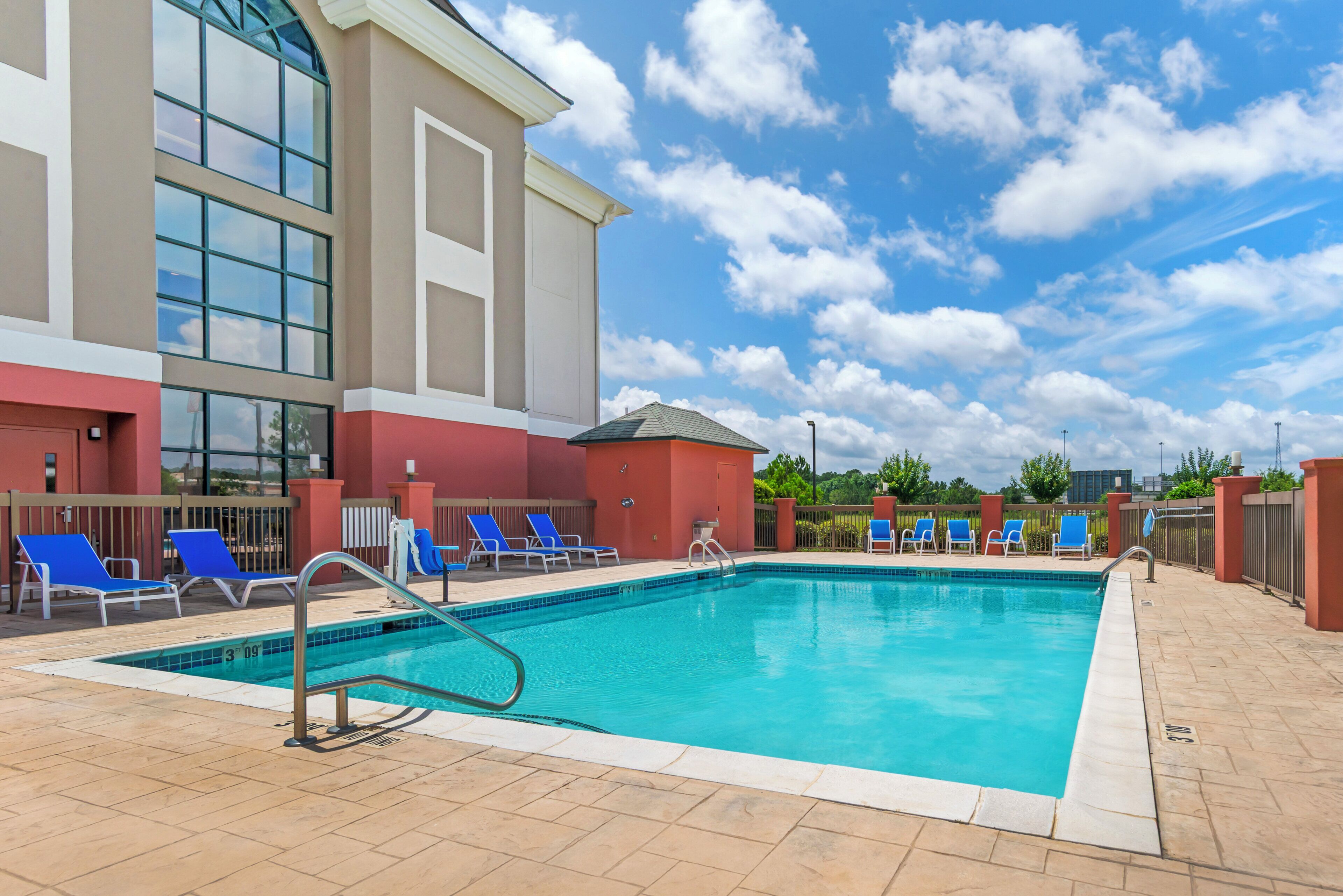A swimming pool with blue lounge chairs, a tiled deck, and a building with large windows and a red facade.