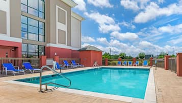 A swimming pool with blue lounge chairs, a tiled deck, and a building with large windows and a red facade.