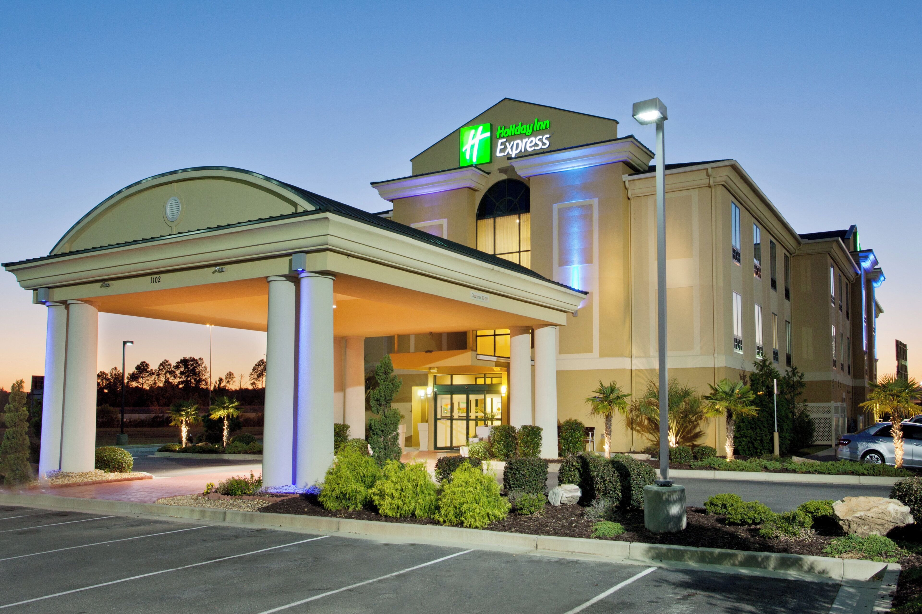 A Holiday Inn Express hotel with a covered entrance, a parking lot, and landscaped areas with shrubs and palm trees.