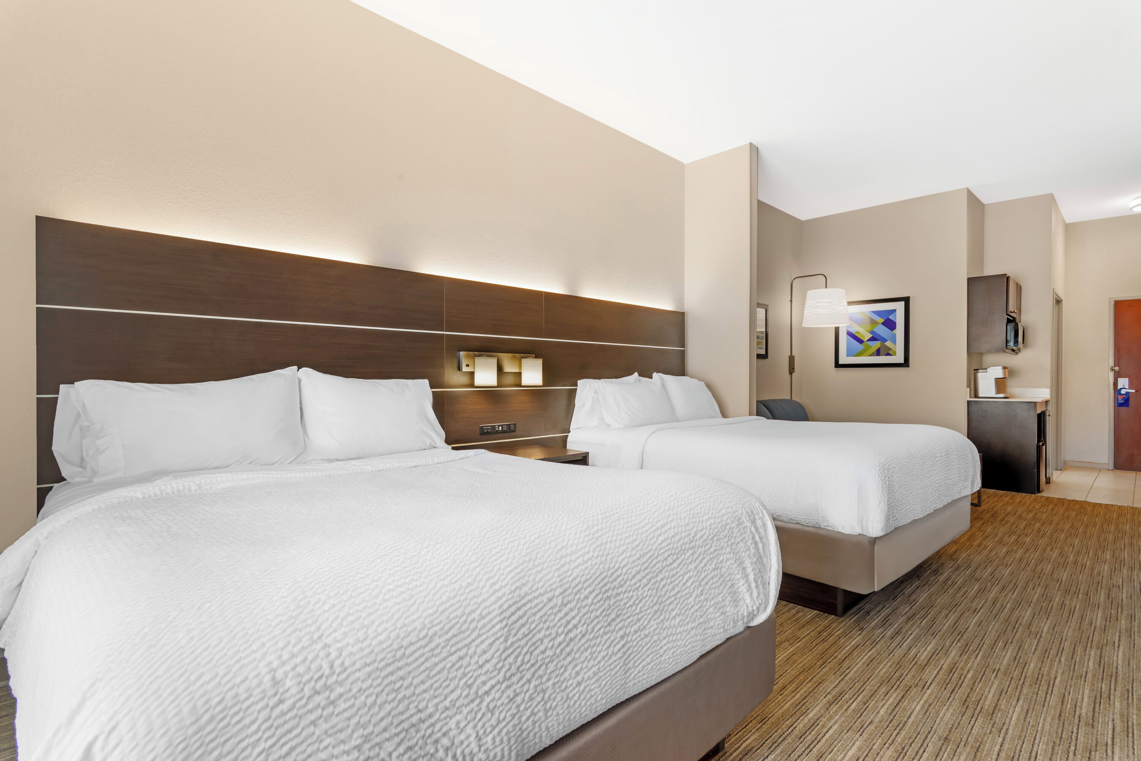 A hotel room with two beds, a nightstand, a lamp, a painting, and a door.