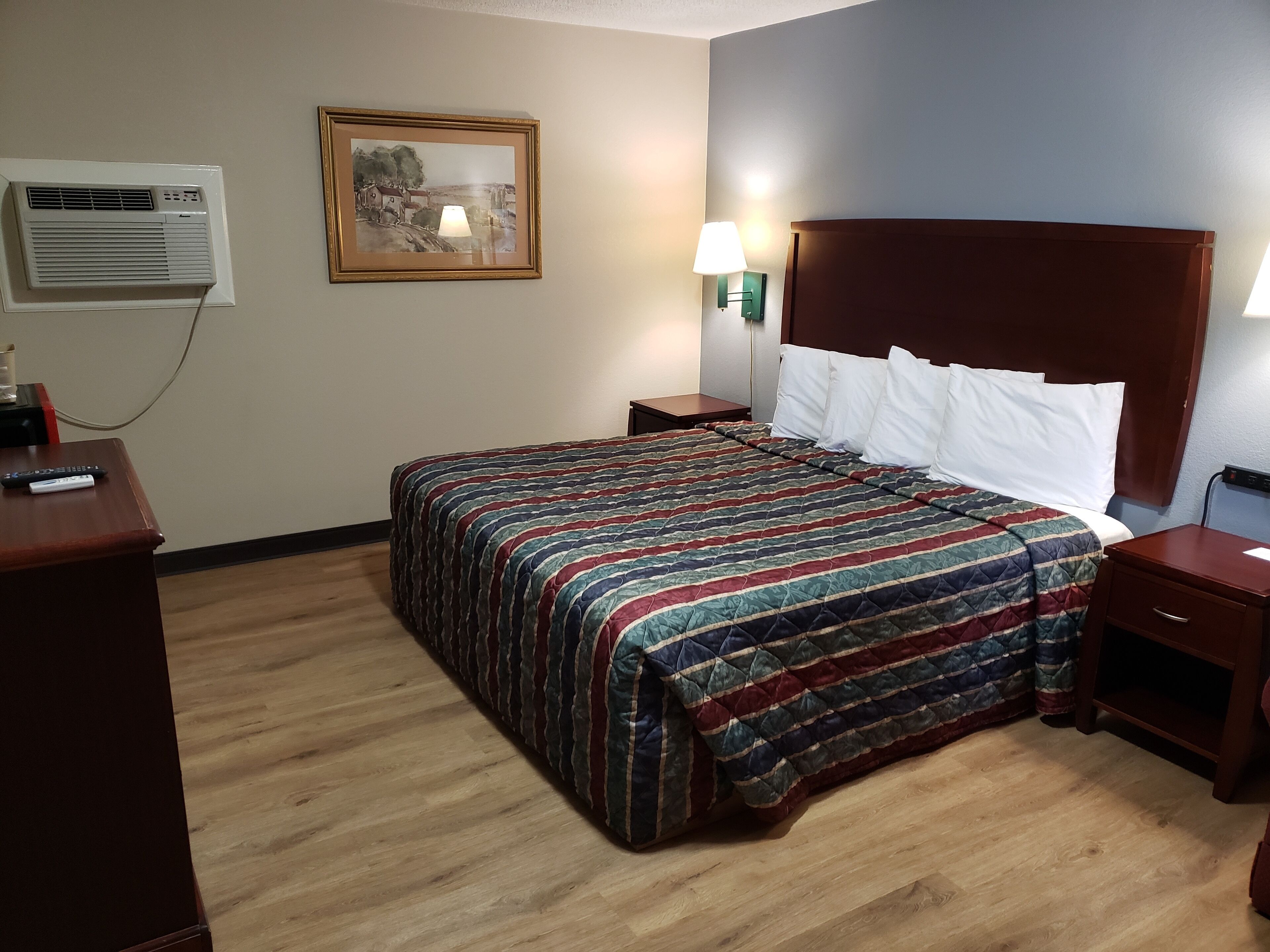 A hotel room with a bed, a nightstand, a lamp, a painting on the wall, and an air conditioning unit.