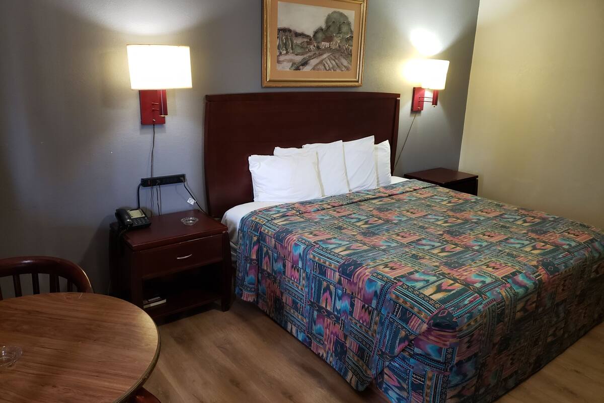 A hotel room with a bed, two bedside lamps, a nightstand with a phone, a wooden chair, and a table.