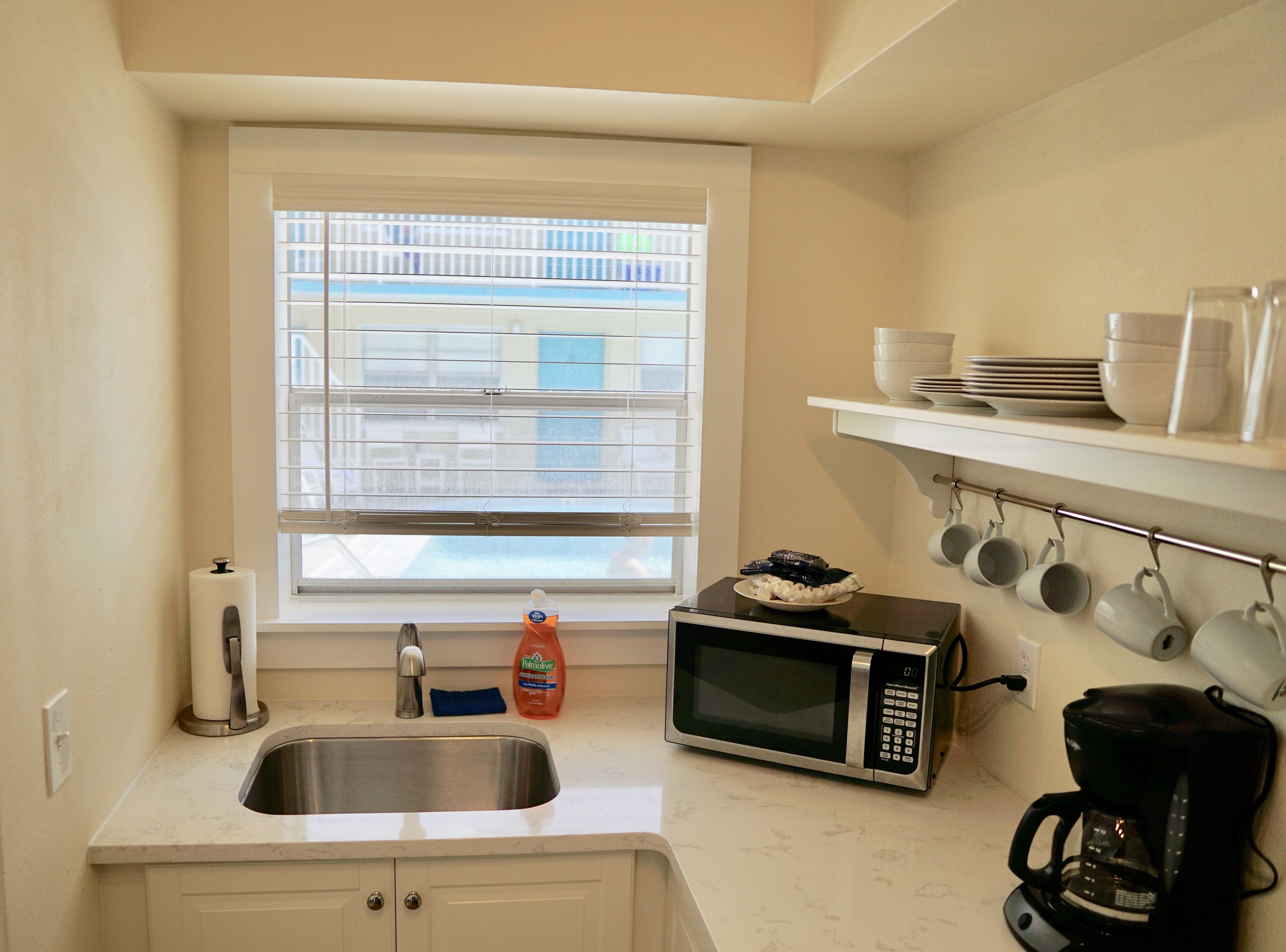 A compact kitchen with a sink, a microwave, a coffee maker, and a window with blinds.
