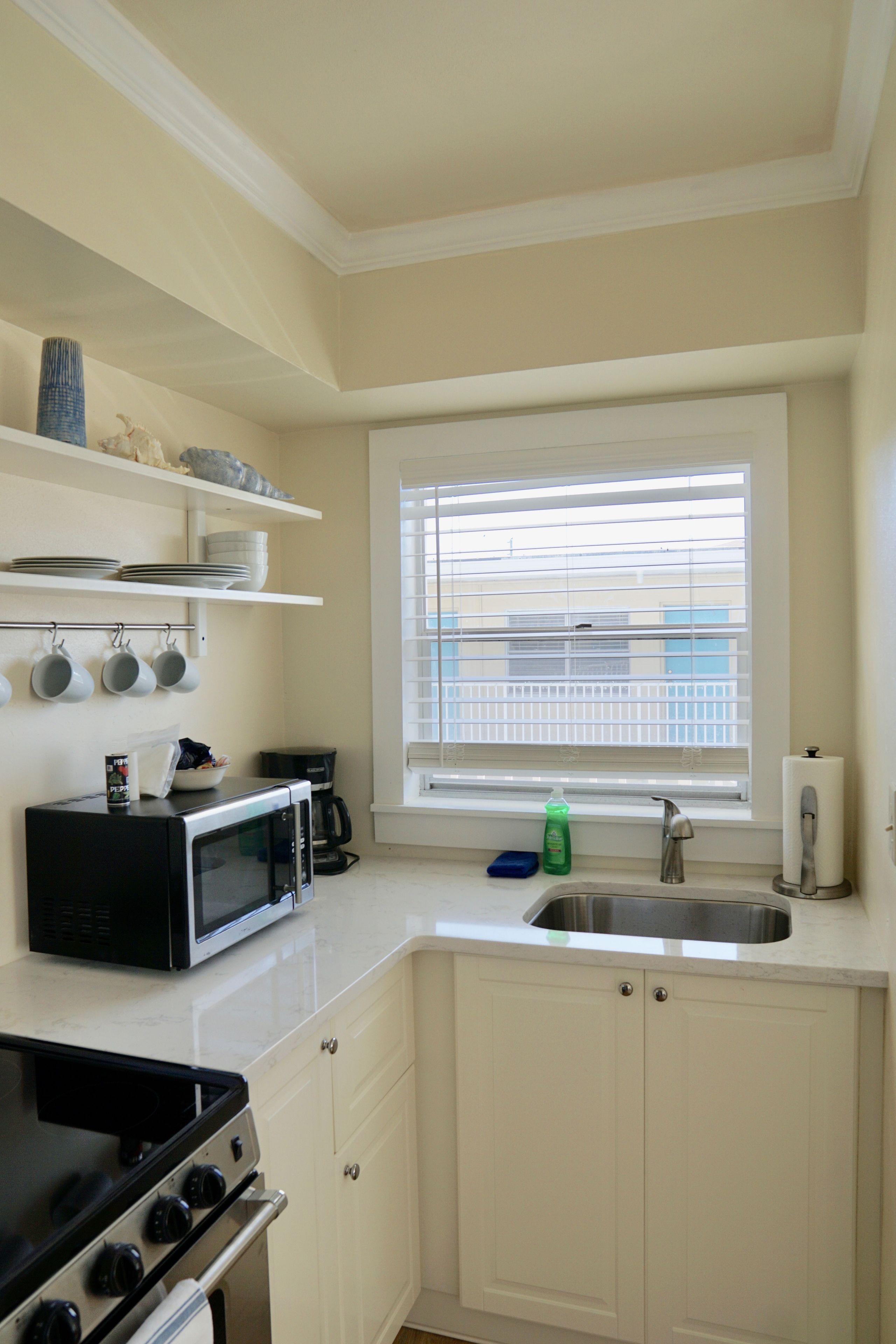 Una cocina con armarios blancos, estufa, microondas, pileta y una ventana con persianas.