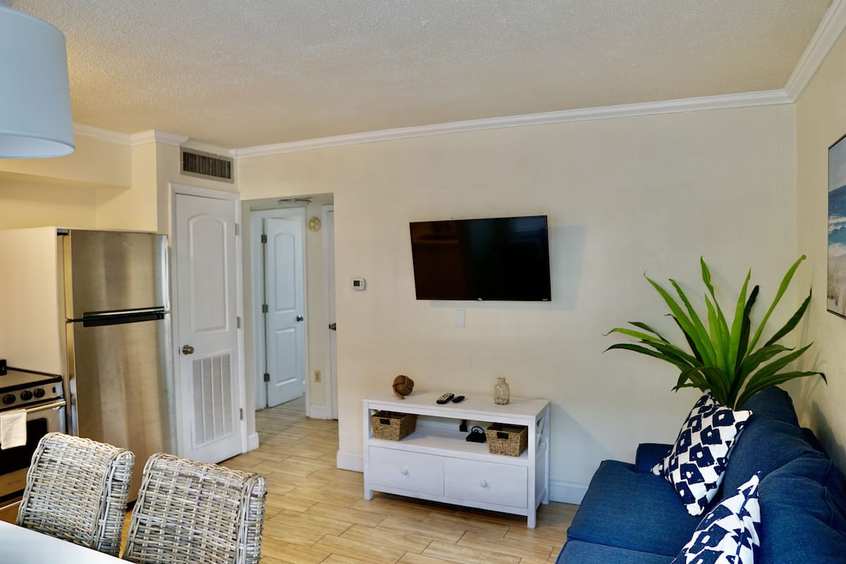 Un salon comprenant un téléviseur, un canapé et des fauteuils en osier. On y trouve une console blanche avec des paniers et une plante.
