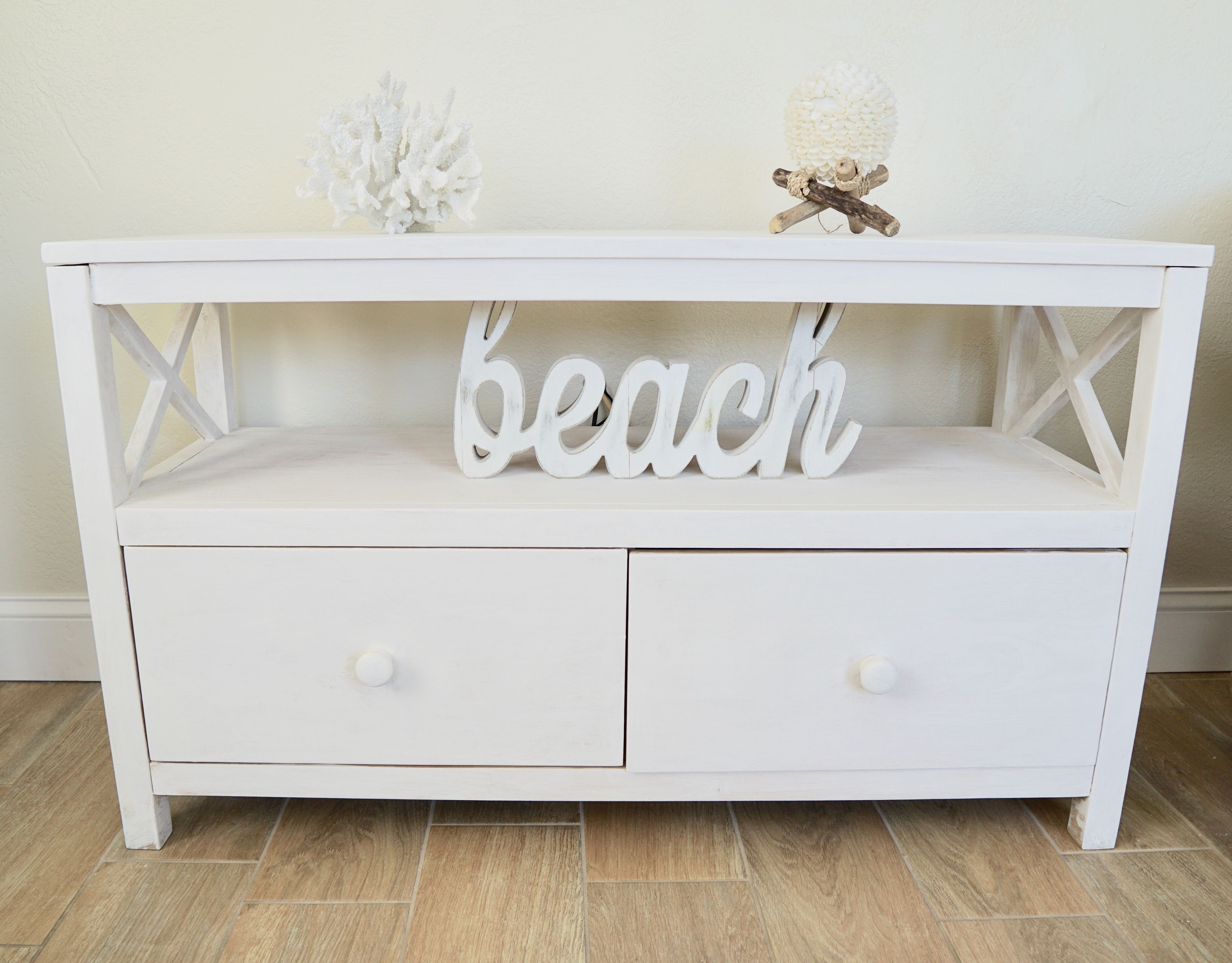 A white wooden console table with two drawers and a shelf. On the shelf, there is a decorative word "beach" and a small white coral-like object.