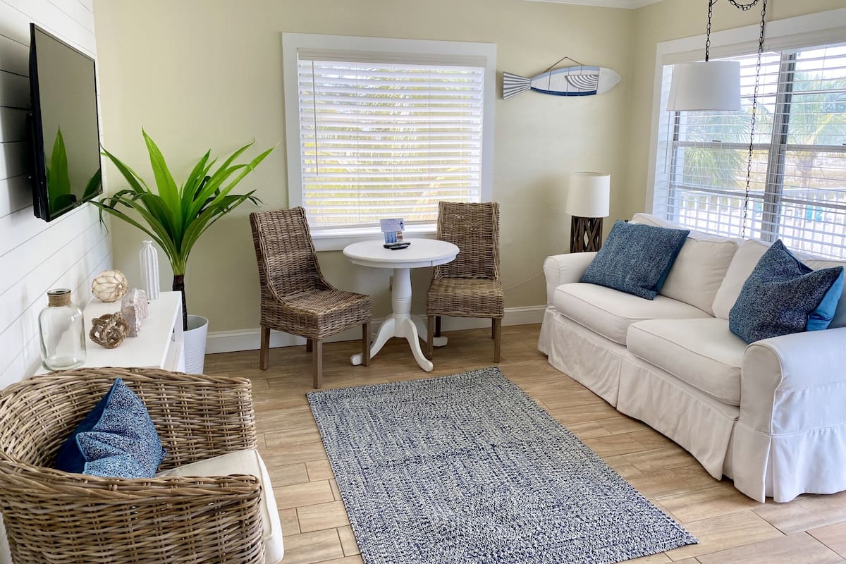 Un salon avec un canapé blanc, deux fauteuils en osier, une table ronde, un tapis bleu et blanc, un téléviseur fixé au mur et un élément de décoration murale en forme de poisson.